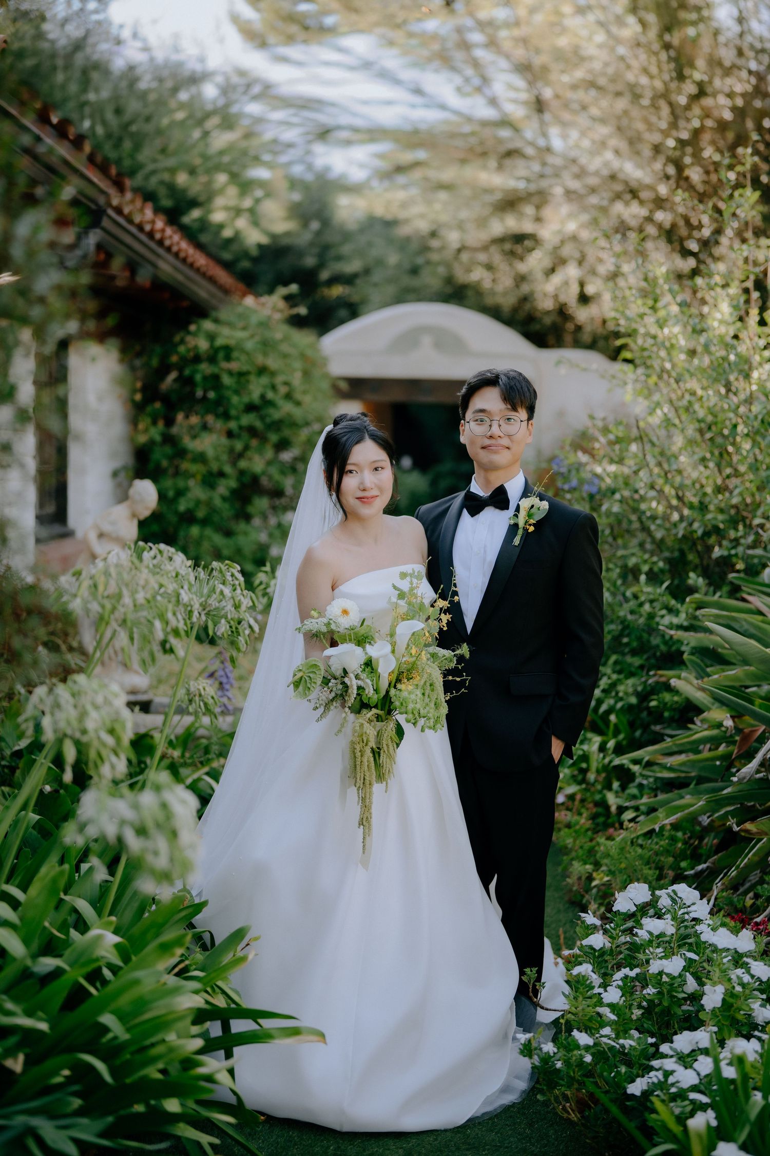 Asian couple posing in a lush garden, bride in strapless gown holding a white bouquet.