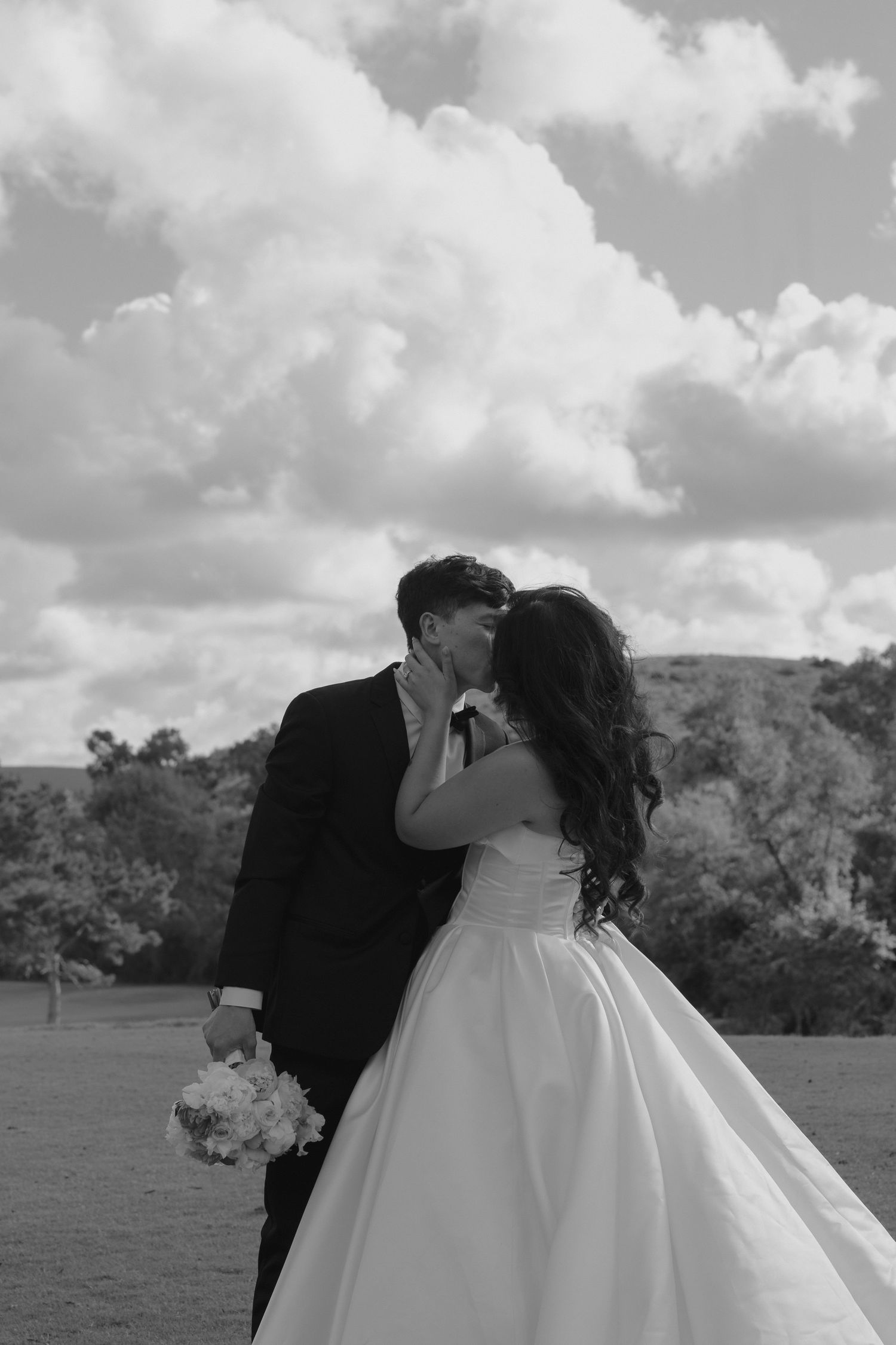 Black and white photo of a bride and groom sharing a kiss outdoors, with a bouquet and cloudy sky backdrop.
