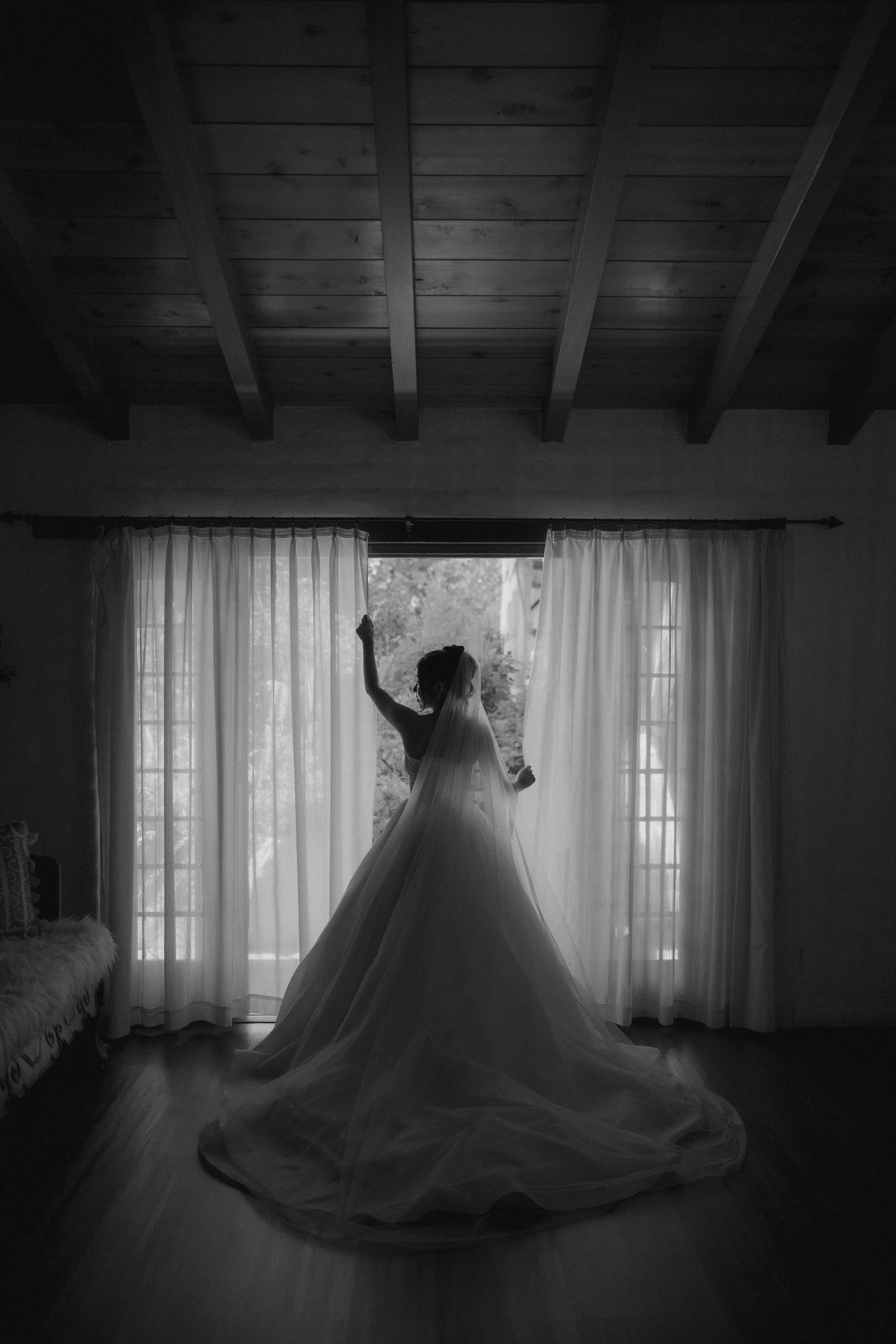 Black and white silhouette of a bride in a ballgown standing before curtained windows in a rustic room.