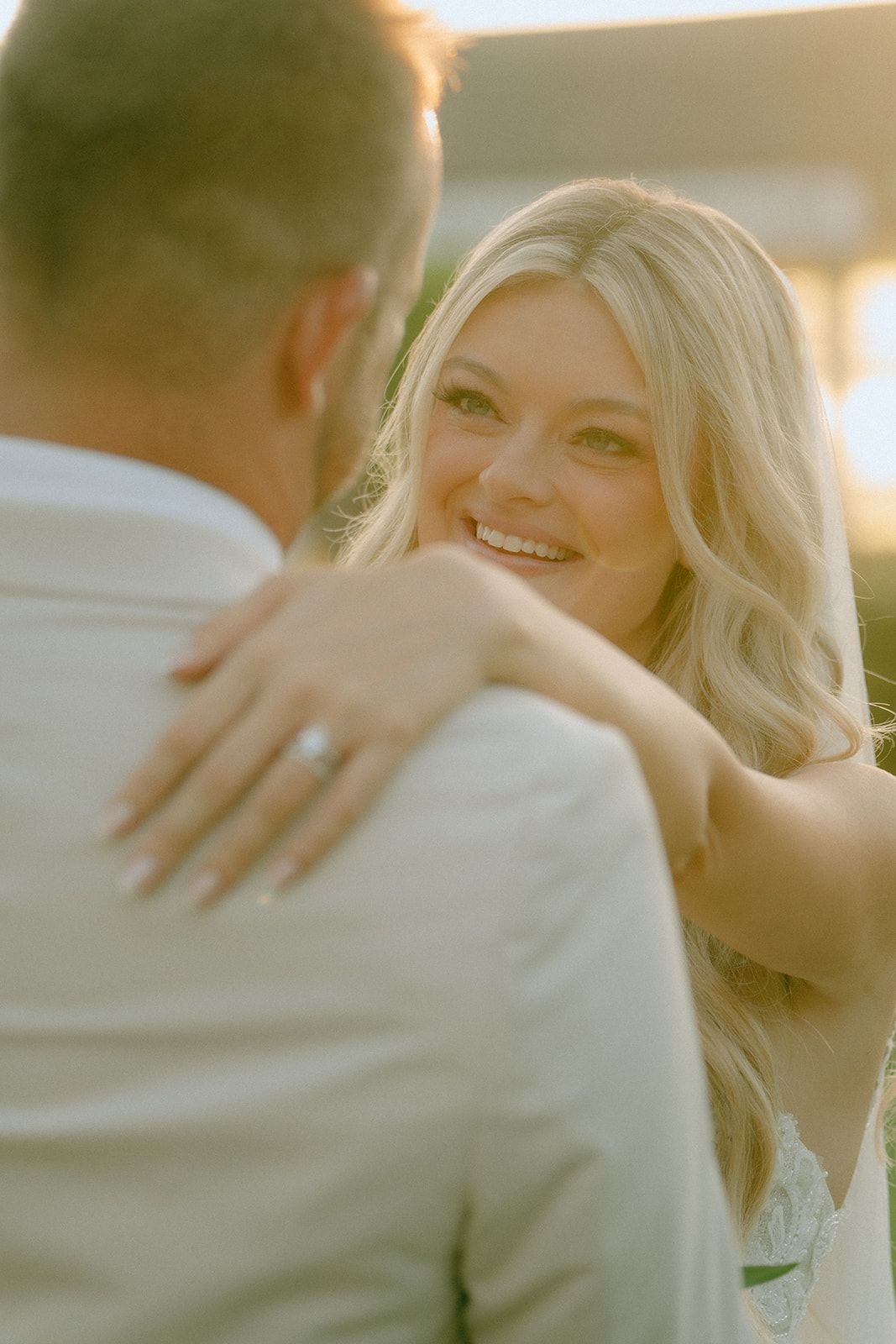 Smiling blonde bride with engagement ring embracing groom in white suit during golden hour wedding photo.