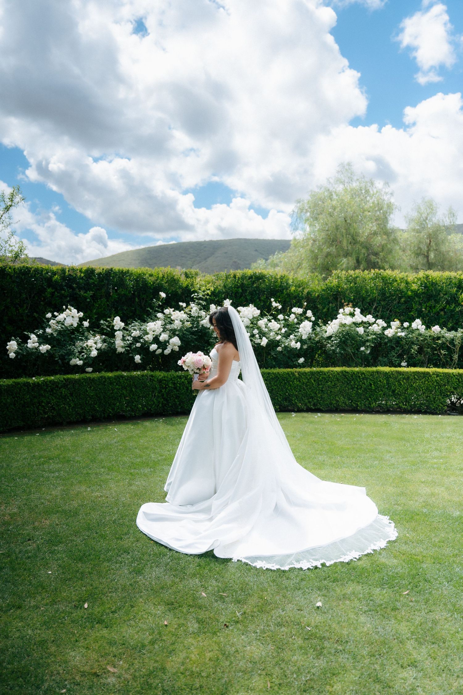 Bride in a white gown with long veil holds pink bouquet in a lush garden with hedges, roses, and mountains.