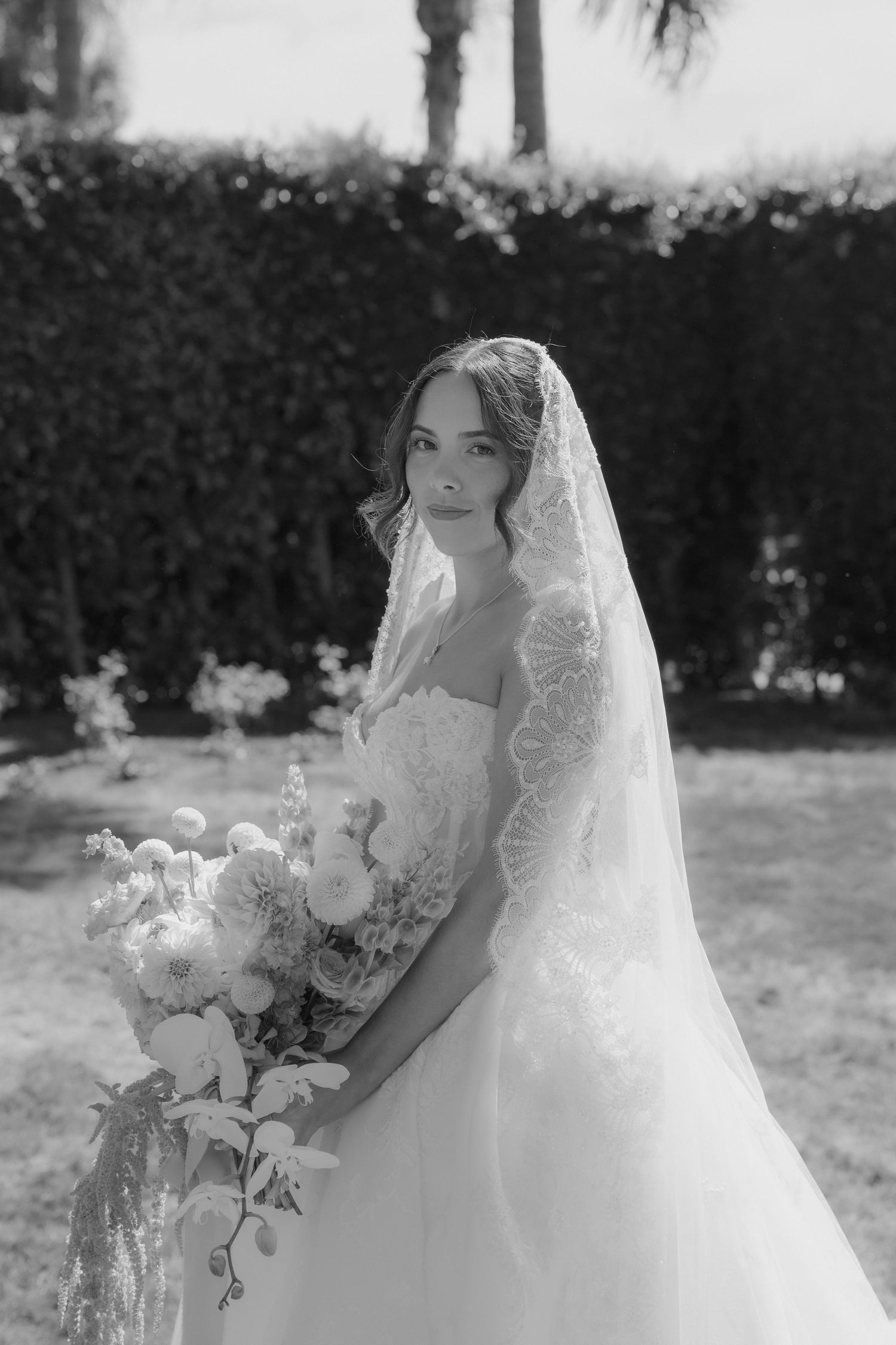 Black and white photo of a bride in a lace veil and strapless gown holding a bouquet outdoors.