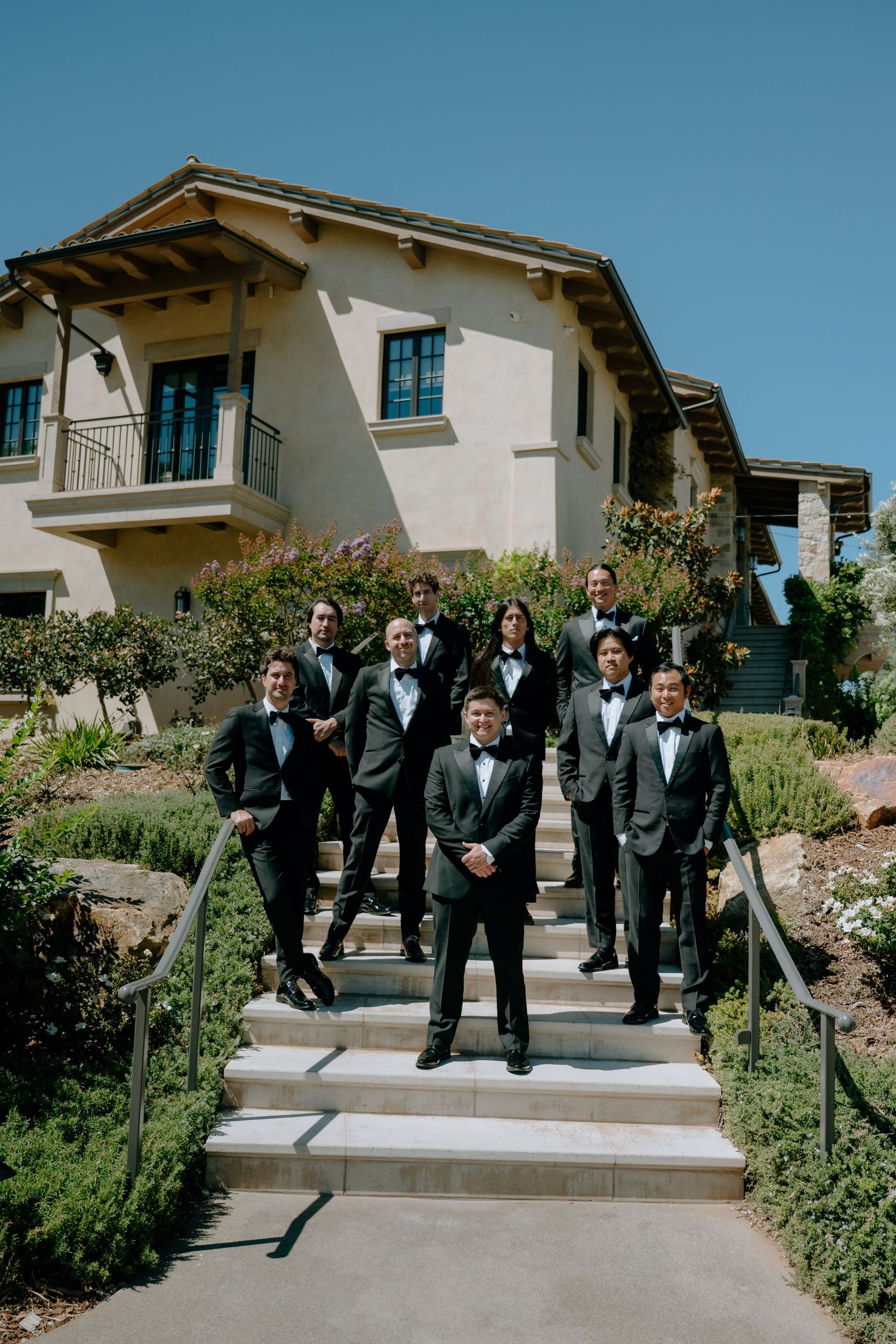 Groom and groomsmen in black tuxedos posing on outdoor steps in front of an elegant venue on a sunny day.