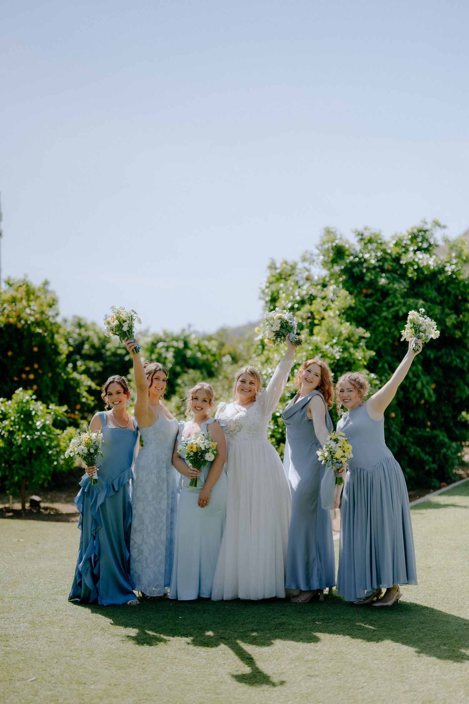 Bride with bridesmaids in blue dresses celebrating joyfully outdoors in sunlight.