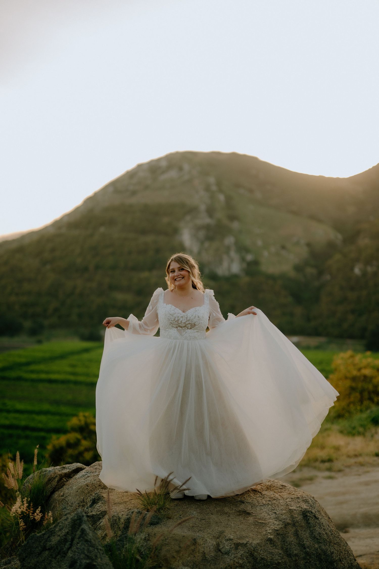 Radiant bride in flowing white gown stands on rocky hilltop with stunning mountain sunset backdrop.