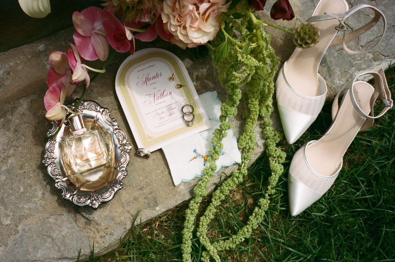 Elegant wedding flat lay with white heels, gold perfume, floral invitation, rings, and green garland on stone.
