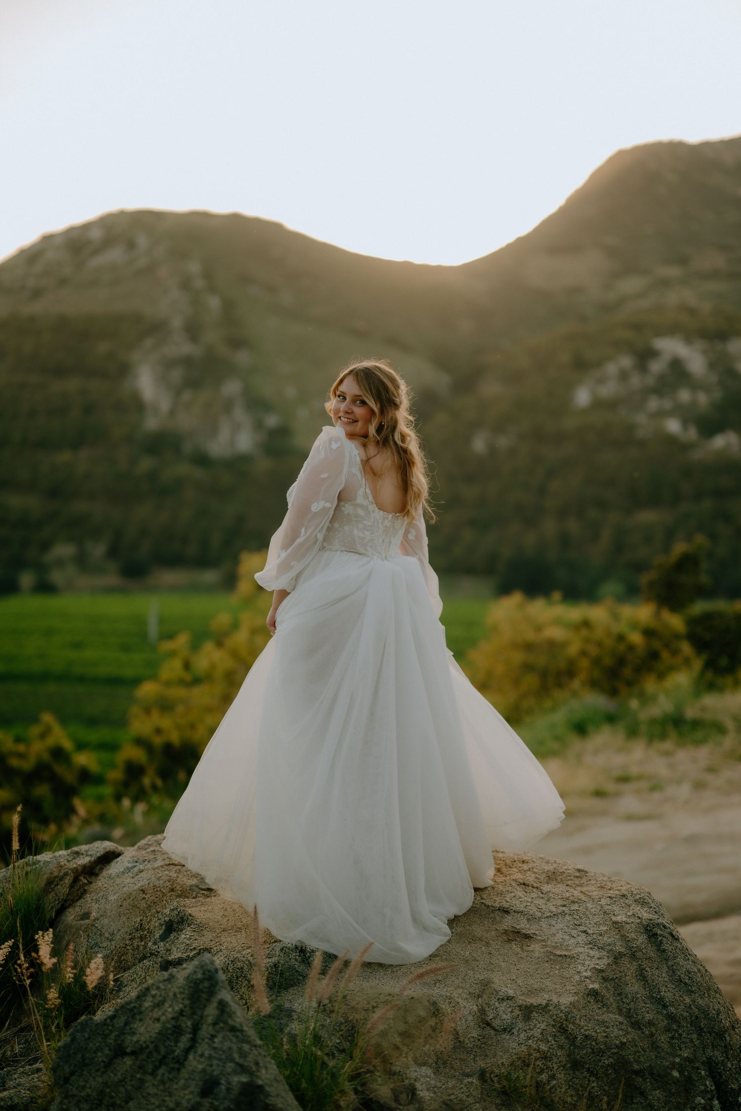 Bride in lace gown standing on a rock at golden sunset with mountain backdrop.