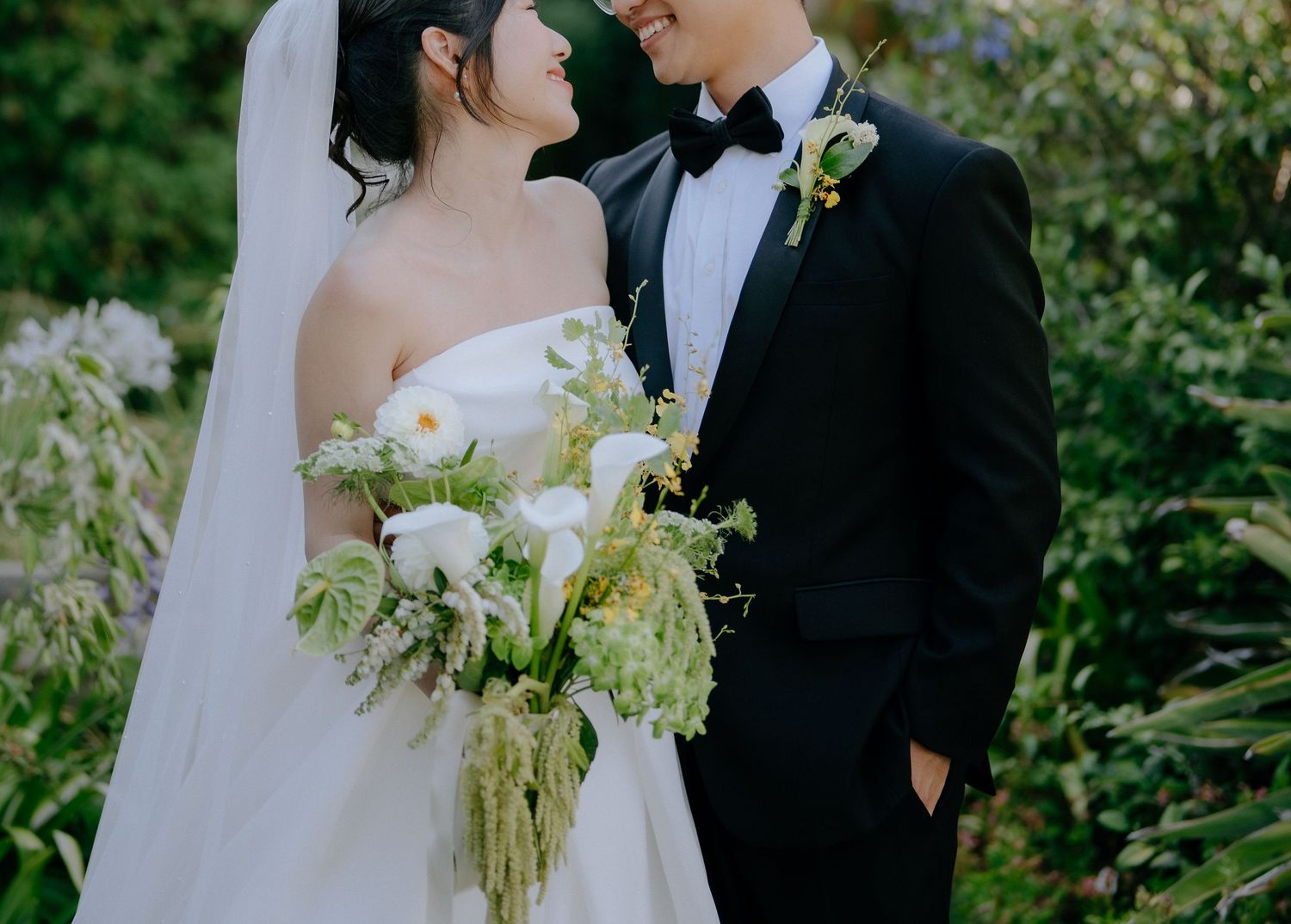 Bride and groom smiling closely, bride holding white calla lily bouquet in lush garden setting.