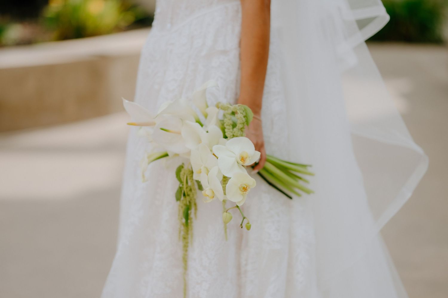 Bride in white lace gown holding elegant white orchid and calla lily wedding bouquet with green stems.