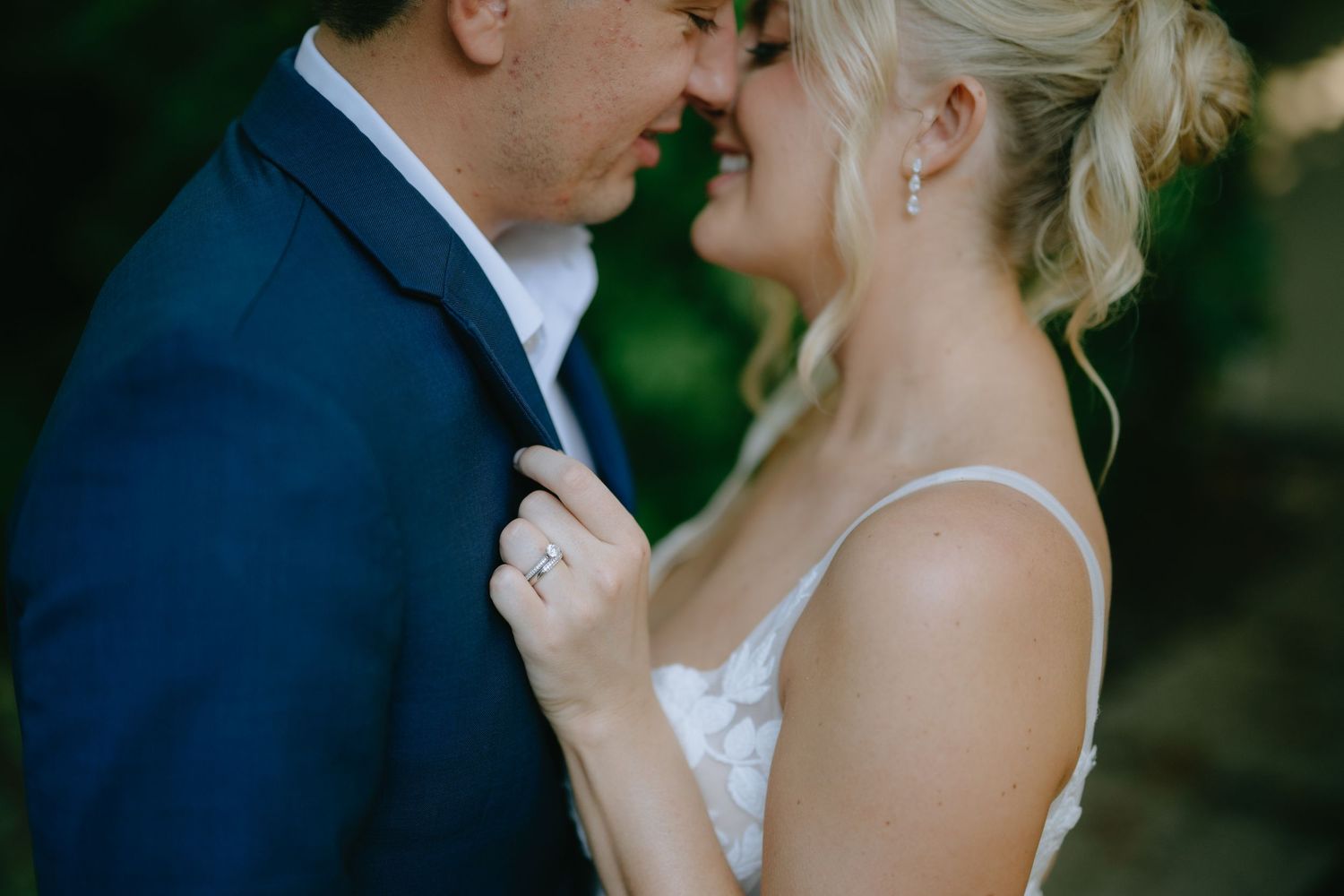 A bride and groom share an intimate moment, touching foreheads, as she holds his lapel wearing a diamond engagement ring.