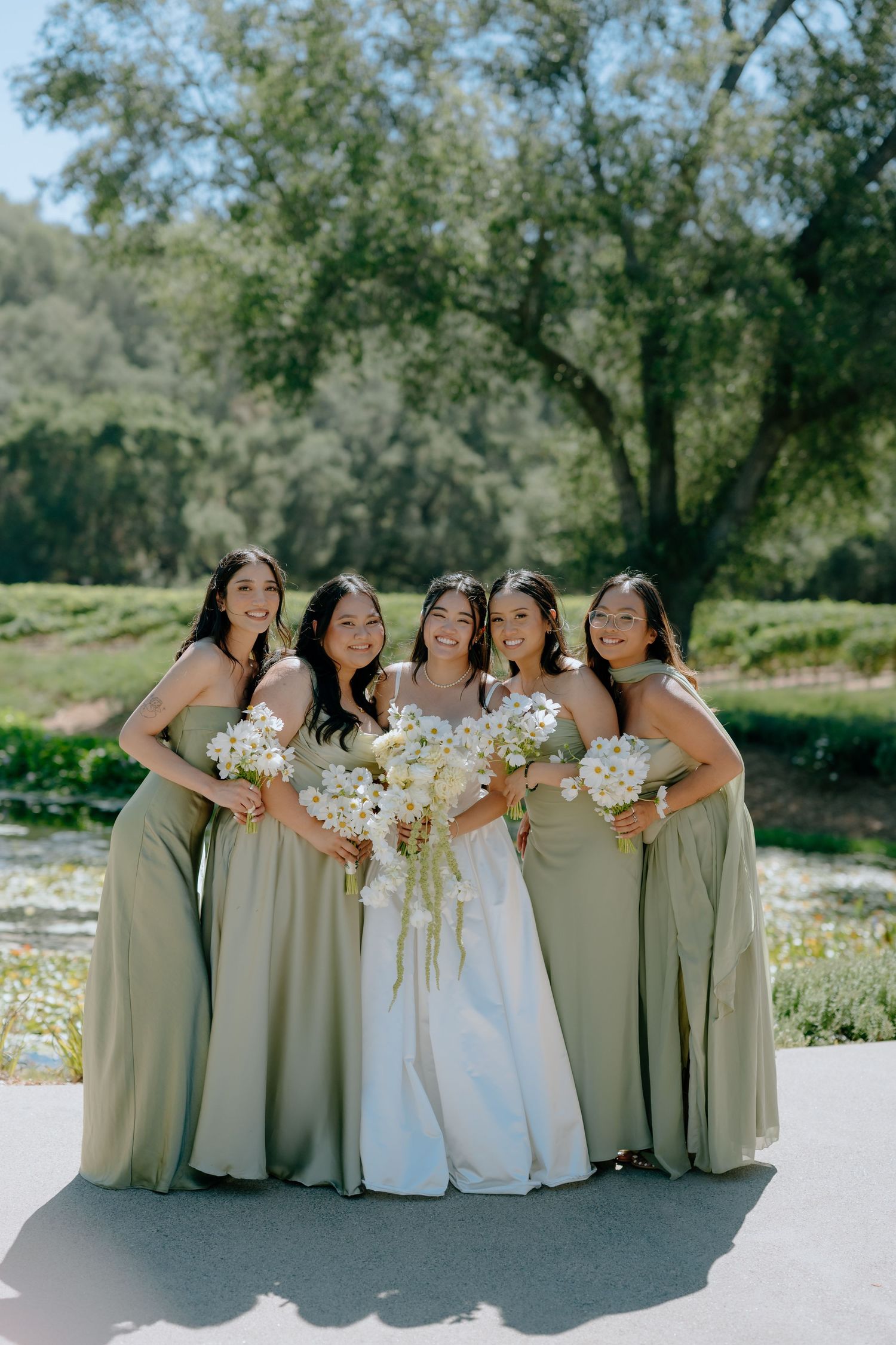 Bride in white gown with four bridesmaids in sage green dresses holding white bouquets outdoors.