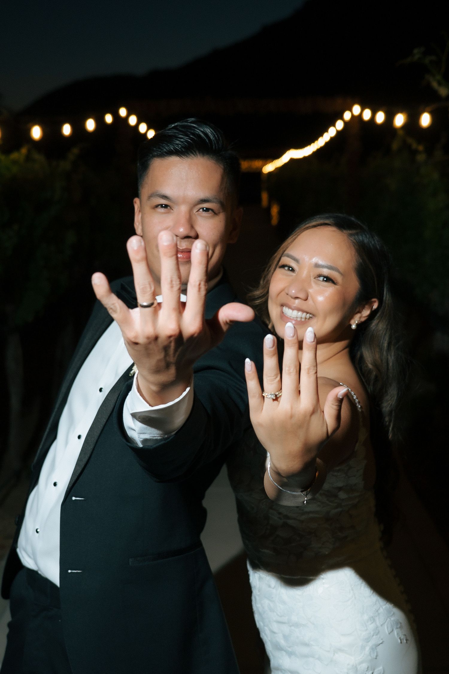 Newlywed couple smiling and showing off their wedding rings at night with string lights in the background.