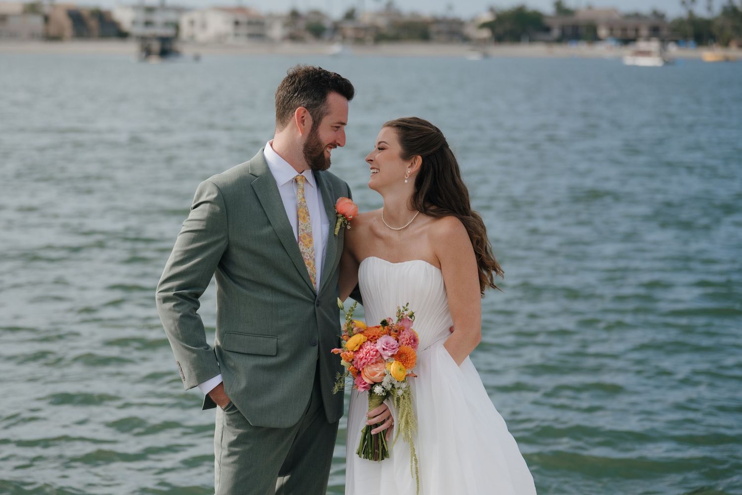 Bride and groom standing by waterfront, bride holding colorful bouquet, groom in gray suit, sharing a romantic moment.