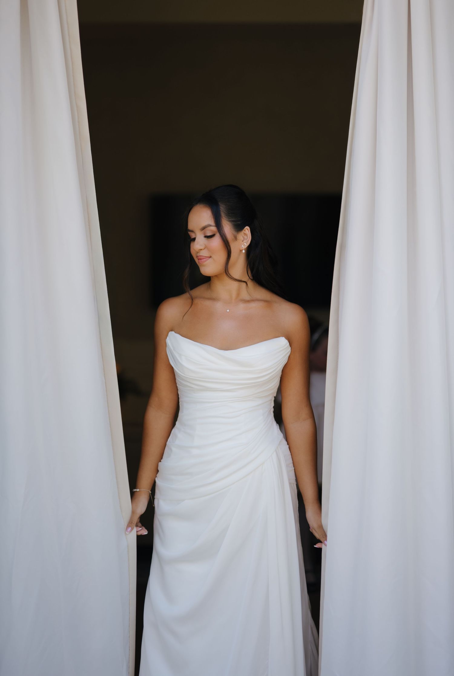 Elegant bride in strapless ruched white gown standing between flowing curtains.