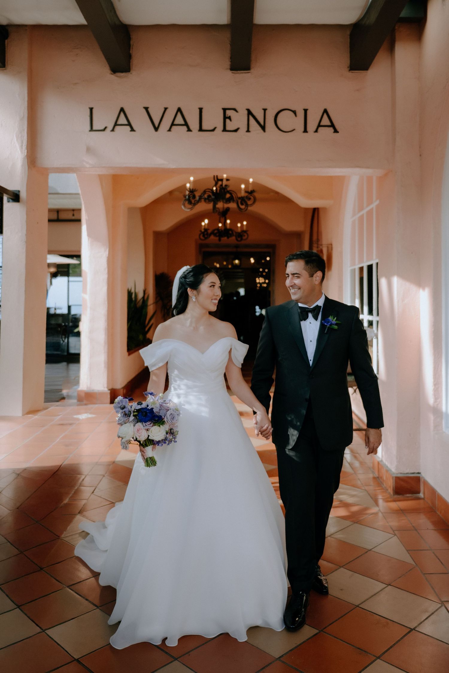 Bride and groom walk hand-in-hand under La Valencia Hotel sign, she holds blue and white bouquet in wedding attire.