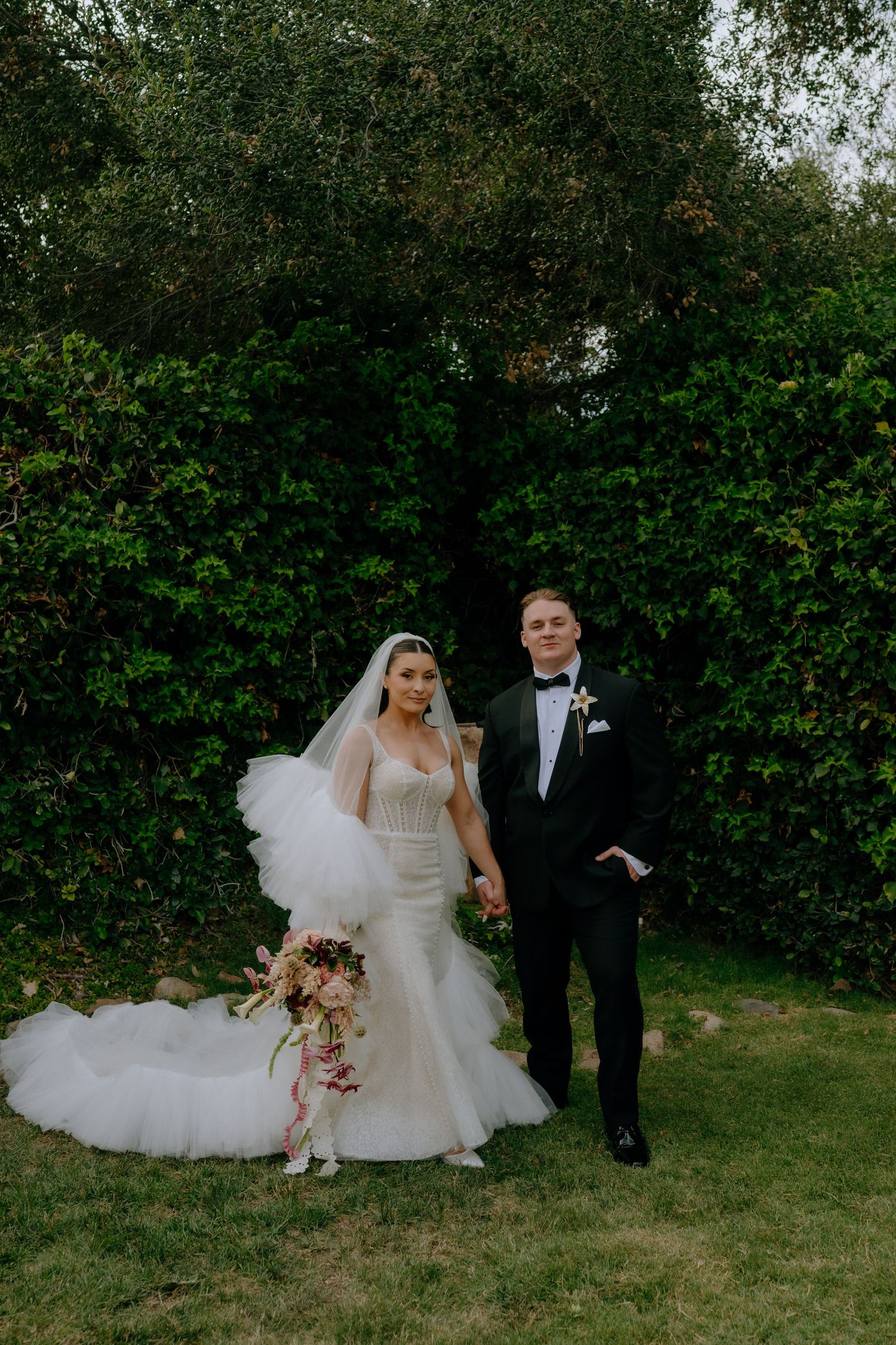 Bride in mermaid gown with veil holds bouquet beside groom in black tuxedo, posing before lush green hedges.