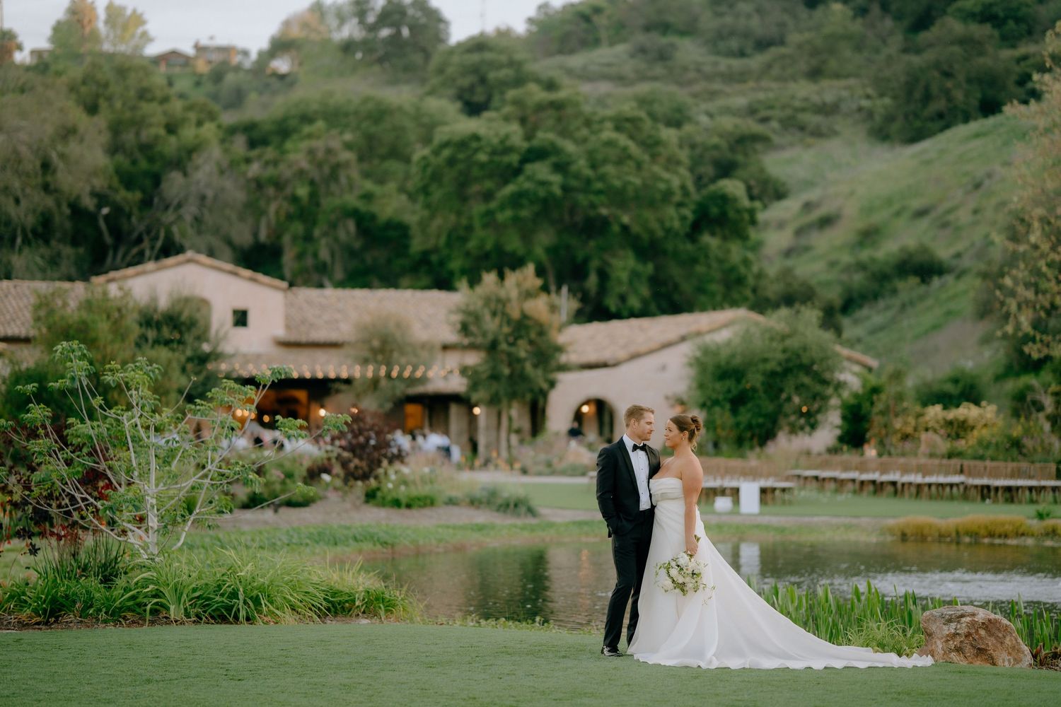 Bride and groom share a romantic moment on lush green lawn with a scenic pond and rustic estate in the background.