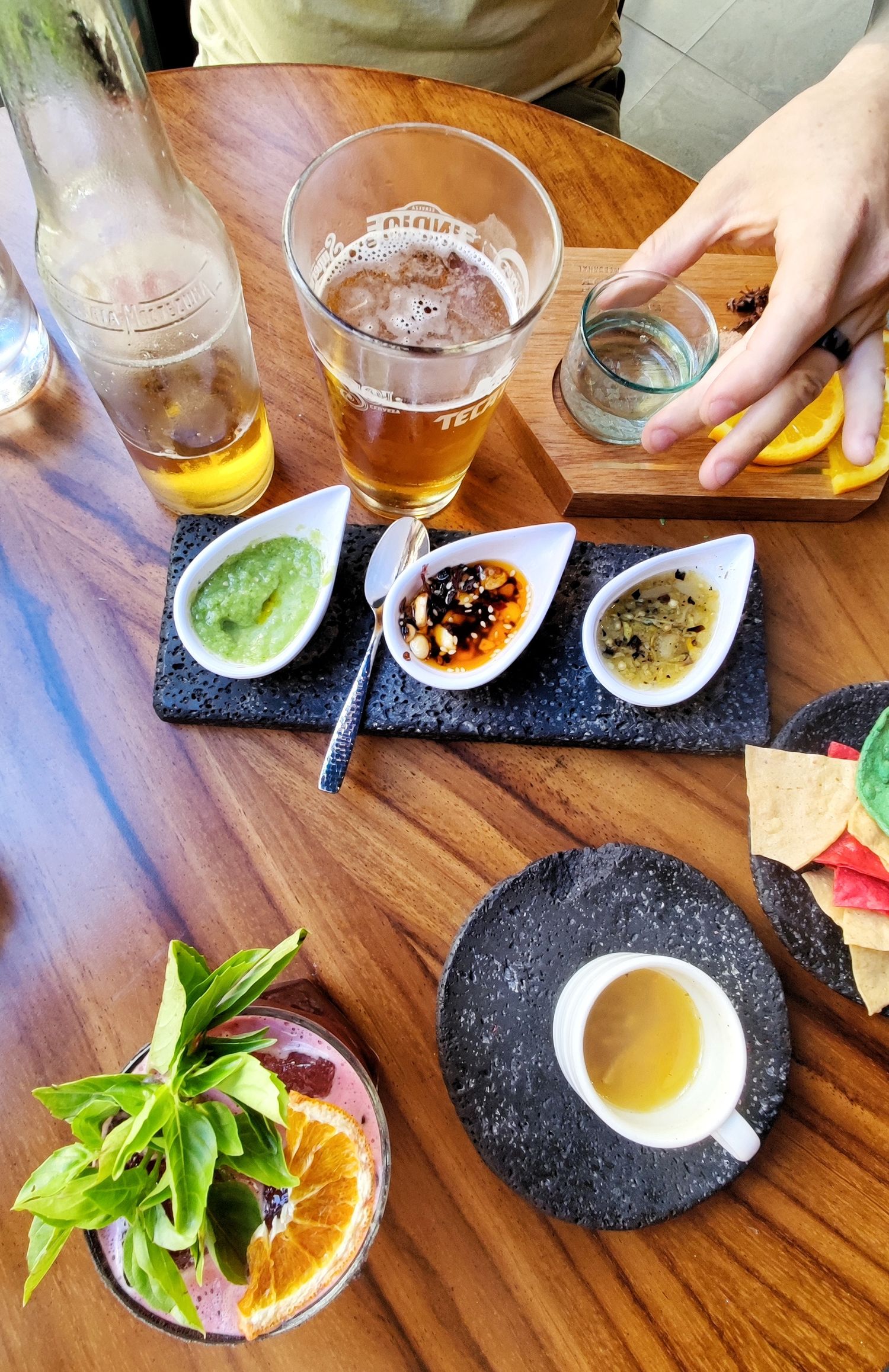 A wooden table is set with three dipping salsas on a slate tray, a cold beer, tortilla chips, a cup of soup and a citrus drink with mint.