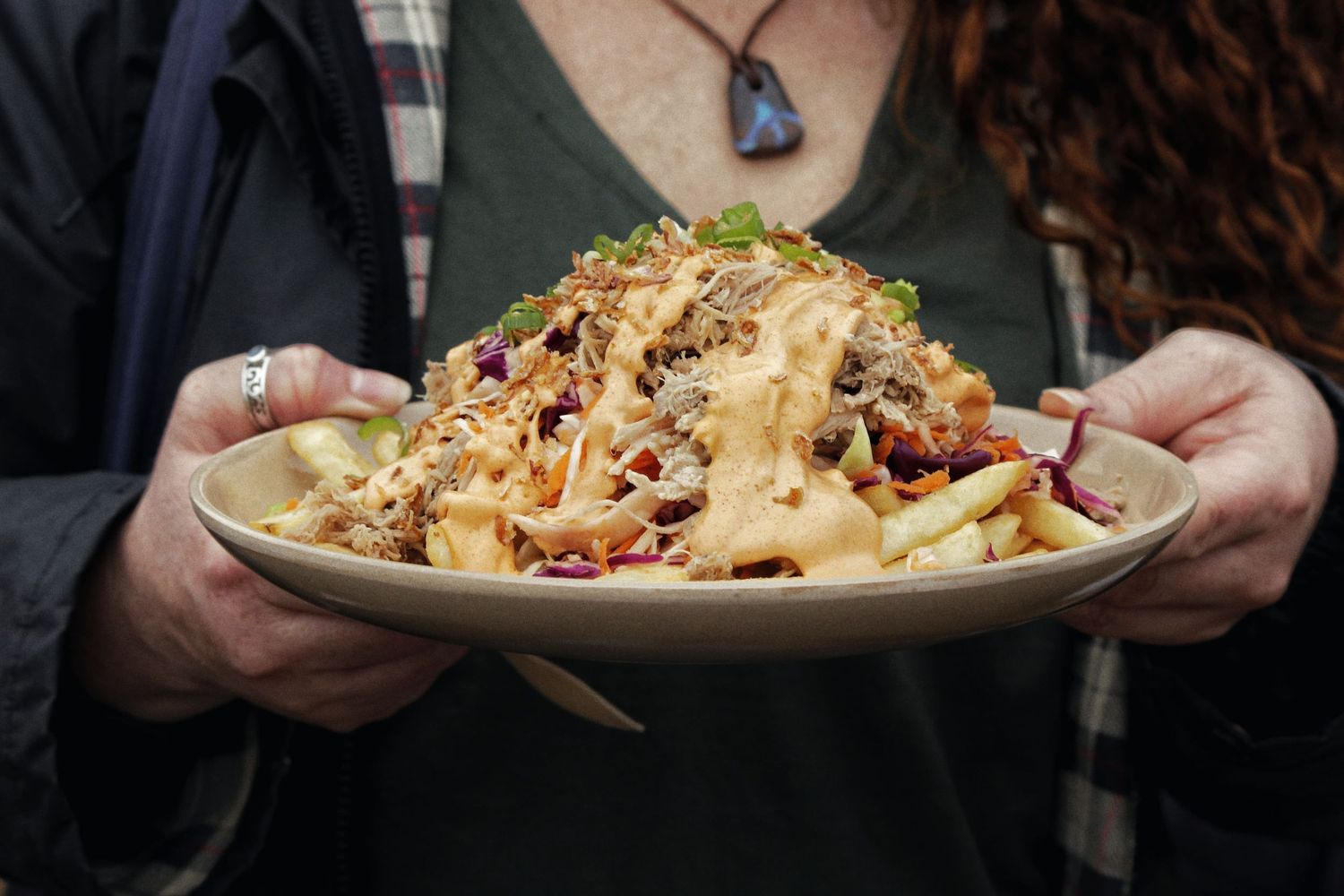 Person in a dark top and pendant necklace holds a plate piled with a fresh slaw salad topped with creamy sauce.
