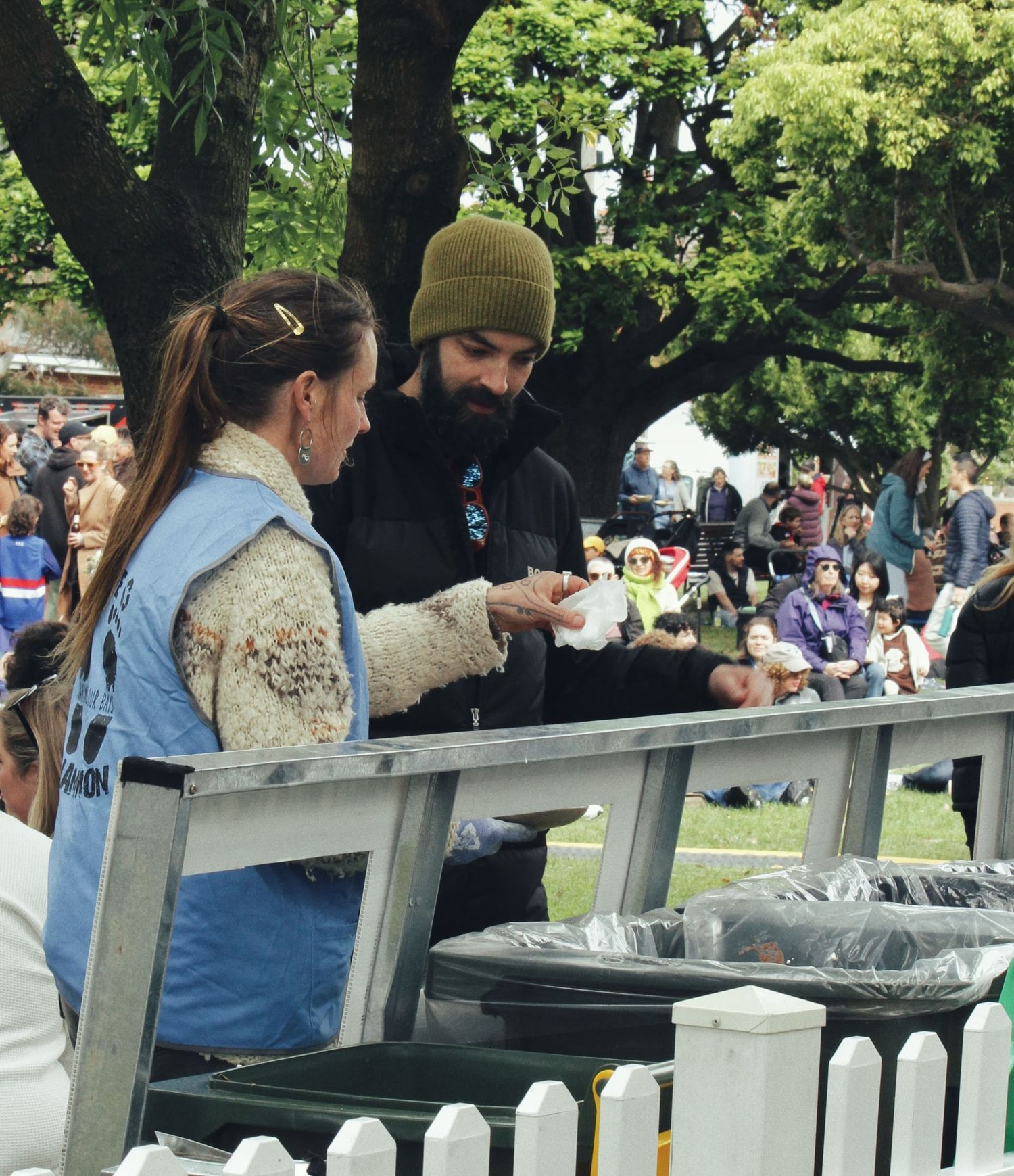 A woman in a blue vest hands food to a bearded man in a beanie at an outdoor festival under large trees.