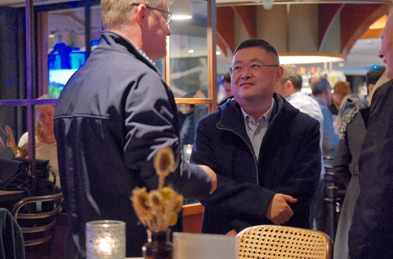 A smiling man in a dark jacket talks with another guest at a lively bar gathering with a candle on the table.