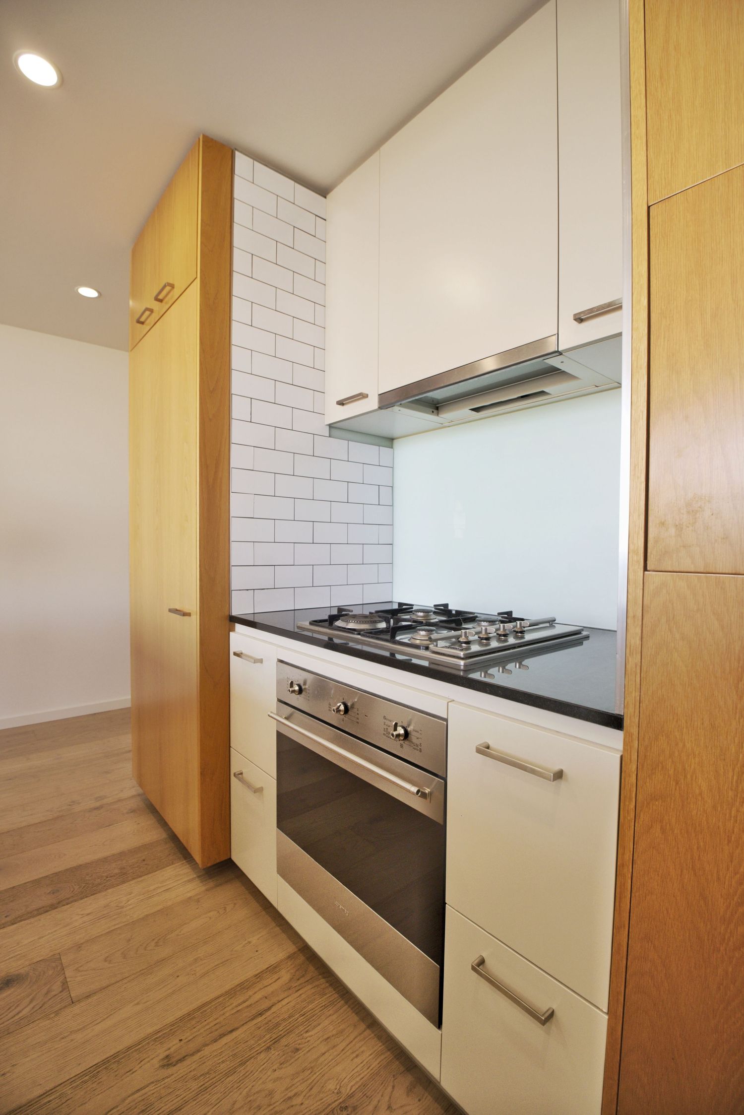 Compact kitchen with gas cooktop, stainless steel oven, white cabinetry, subway tile splashback and timber joinery.