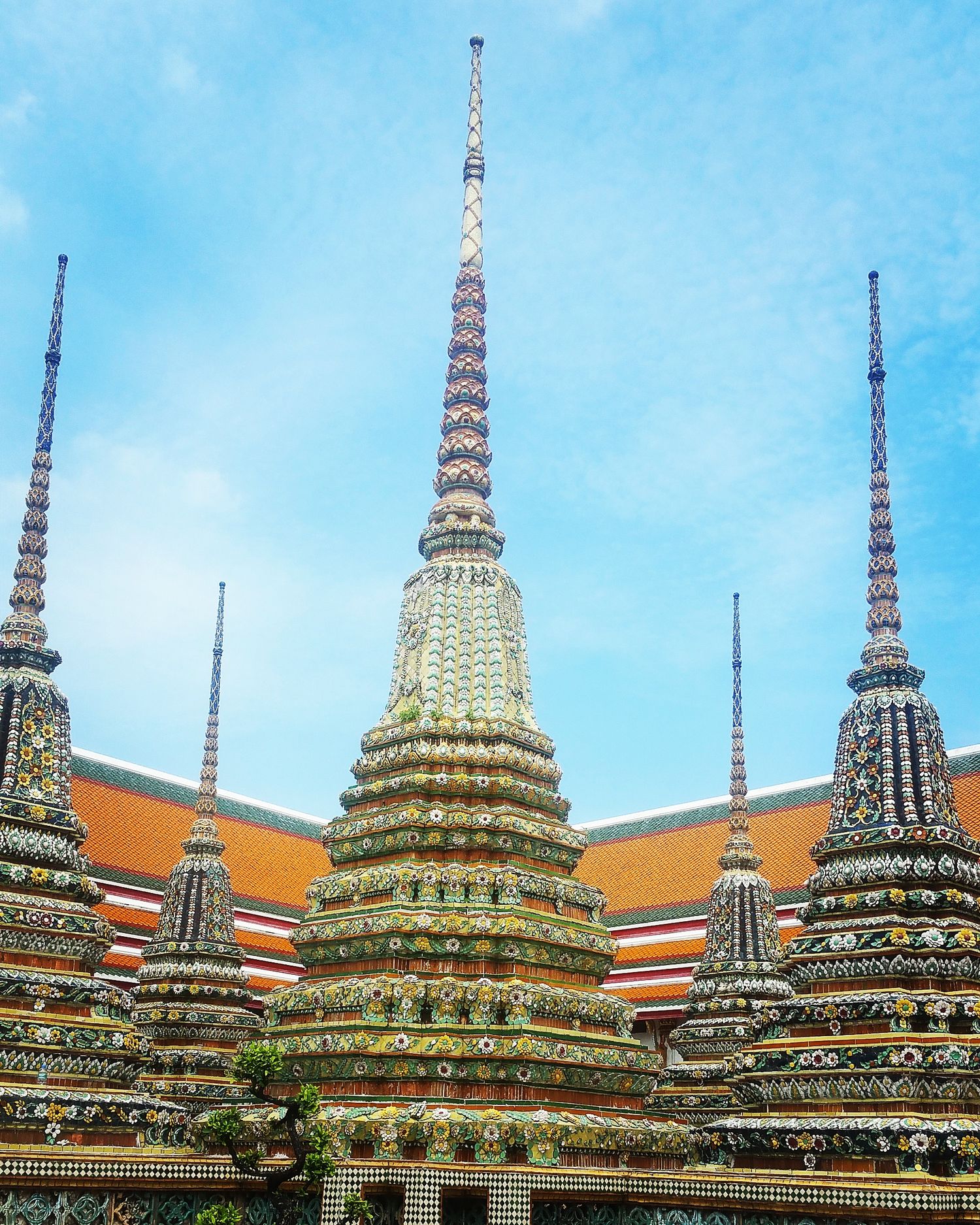 Tall decorated pagodas covered in colorful ceramic mosaic tiles beside orange temple roofs under a bright blue sky in Bangkok.
