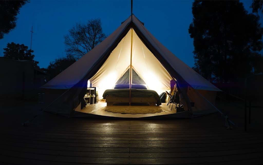 Glowing bell tent illuminated at dusk on a wooden deck, with a cozy bed visible inside and trees in the background.