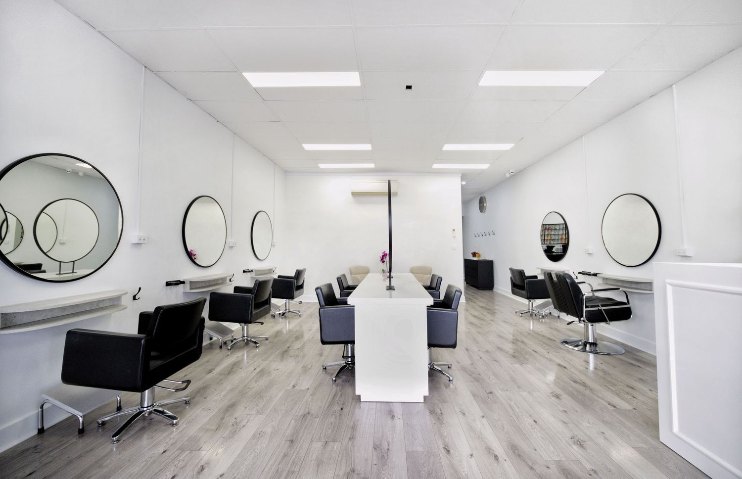 Bright modern hair salon interior with black styling chairs, round wall mirrors, a central white table and light timber flooring.