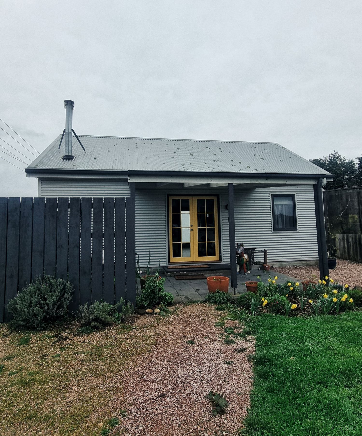 A small grey weatherboard cottage with yellow double doors, a metal chimney flue and daffodils in the front garden.