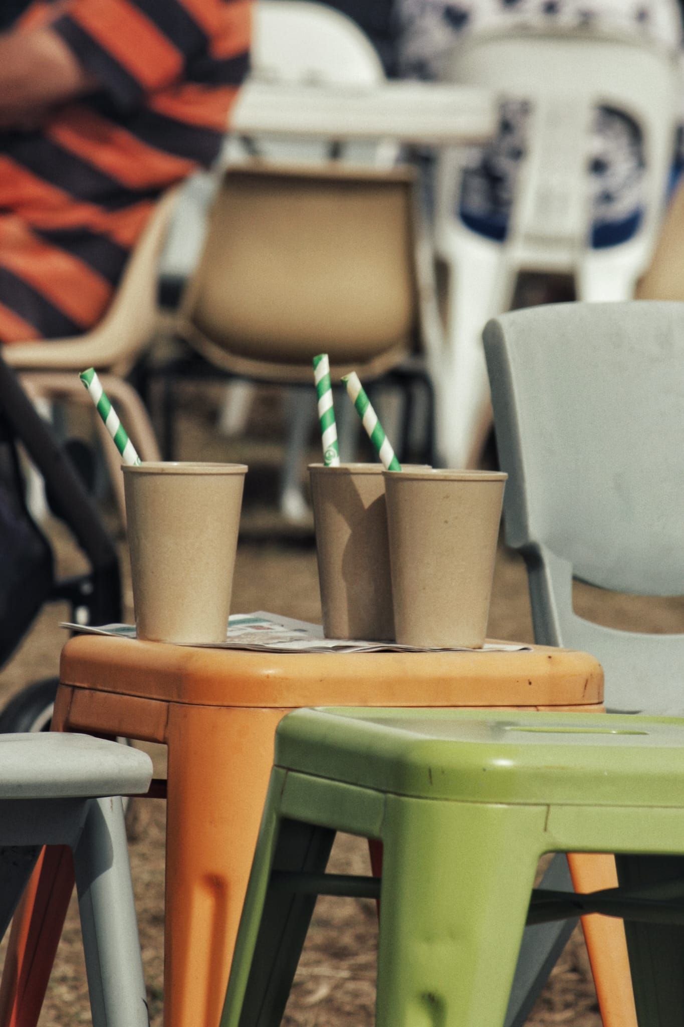 Three paper cups with green striped paper straws rest on an orange metal stool beside colorful chairs at a festival.