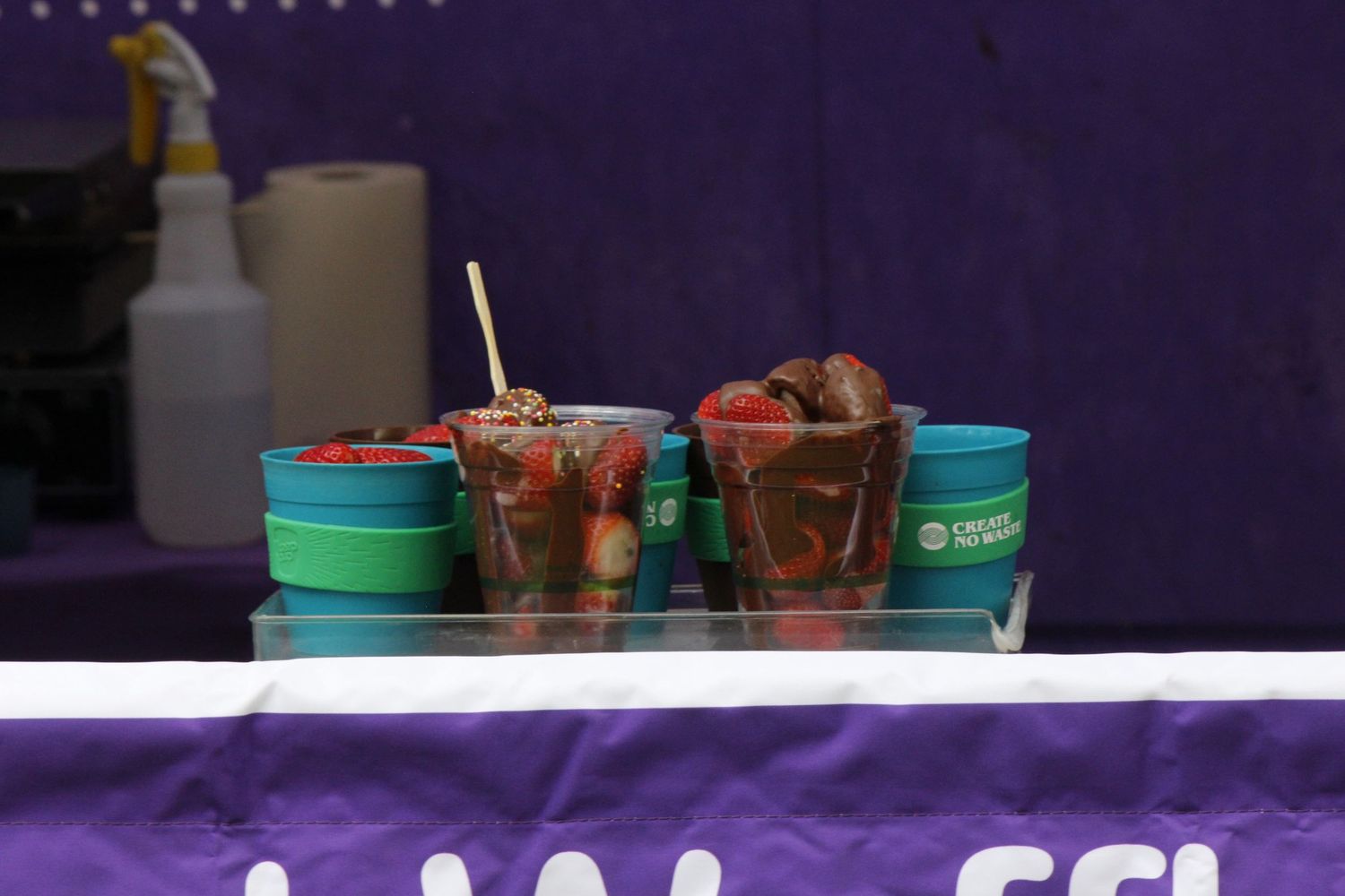 Two plastic cups filled with chocolate-dipped strawberries sit on a purple-draped counter at a festival food stall.