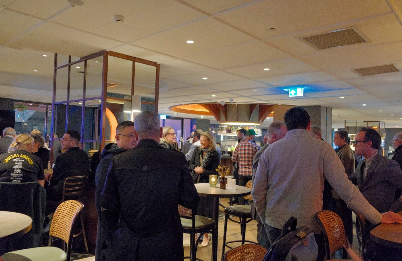 A crowded bar interior with people standing and chatting around high tables under bright ceiling lights during a social event.