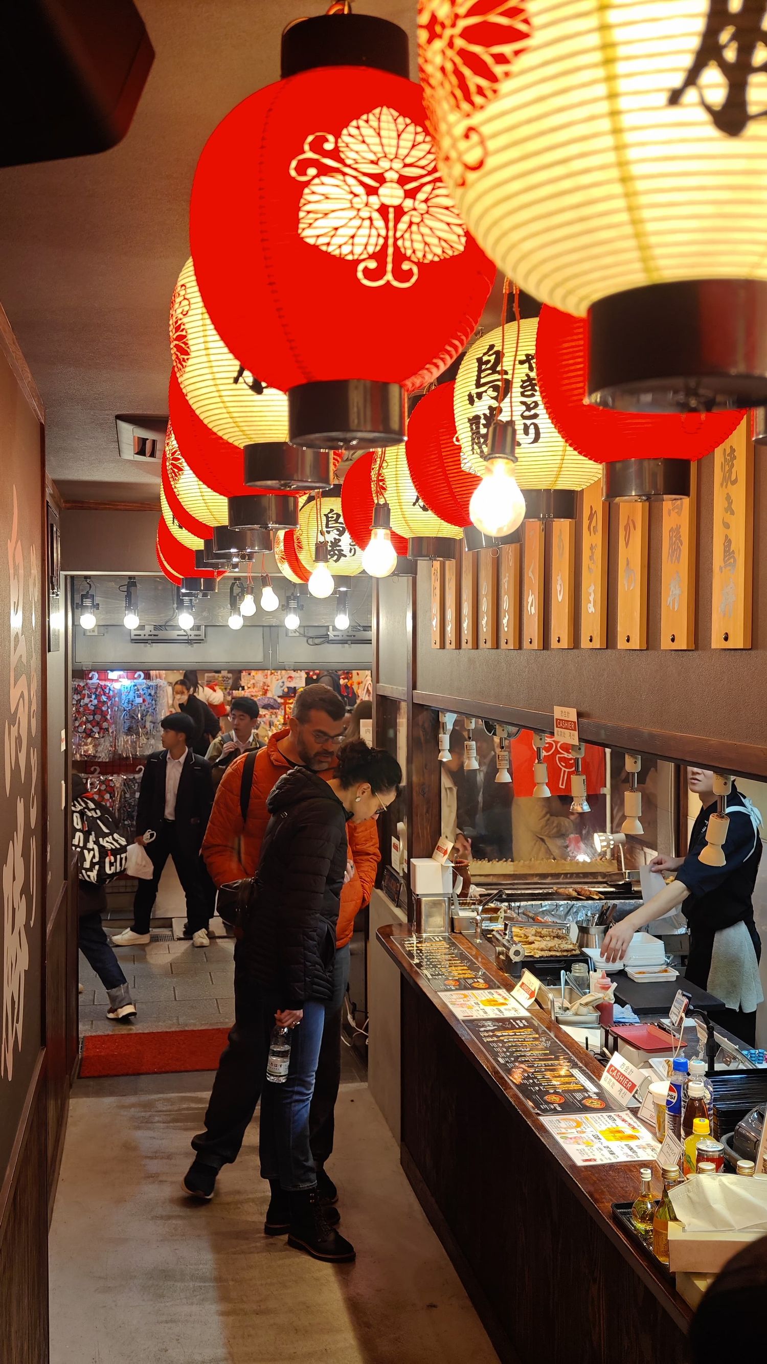 Red paper lanterns hang above a lively Japanese street food alley as visitors browse stalls and grilled snacks.