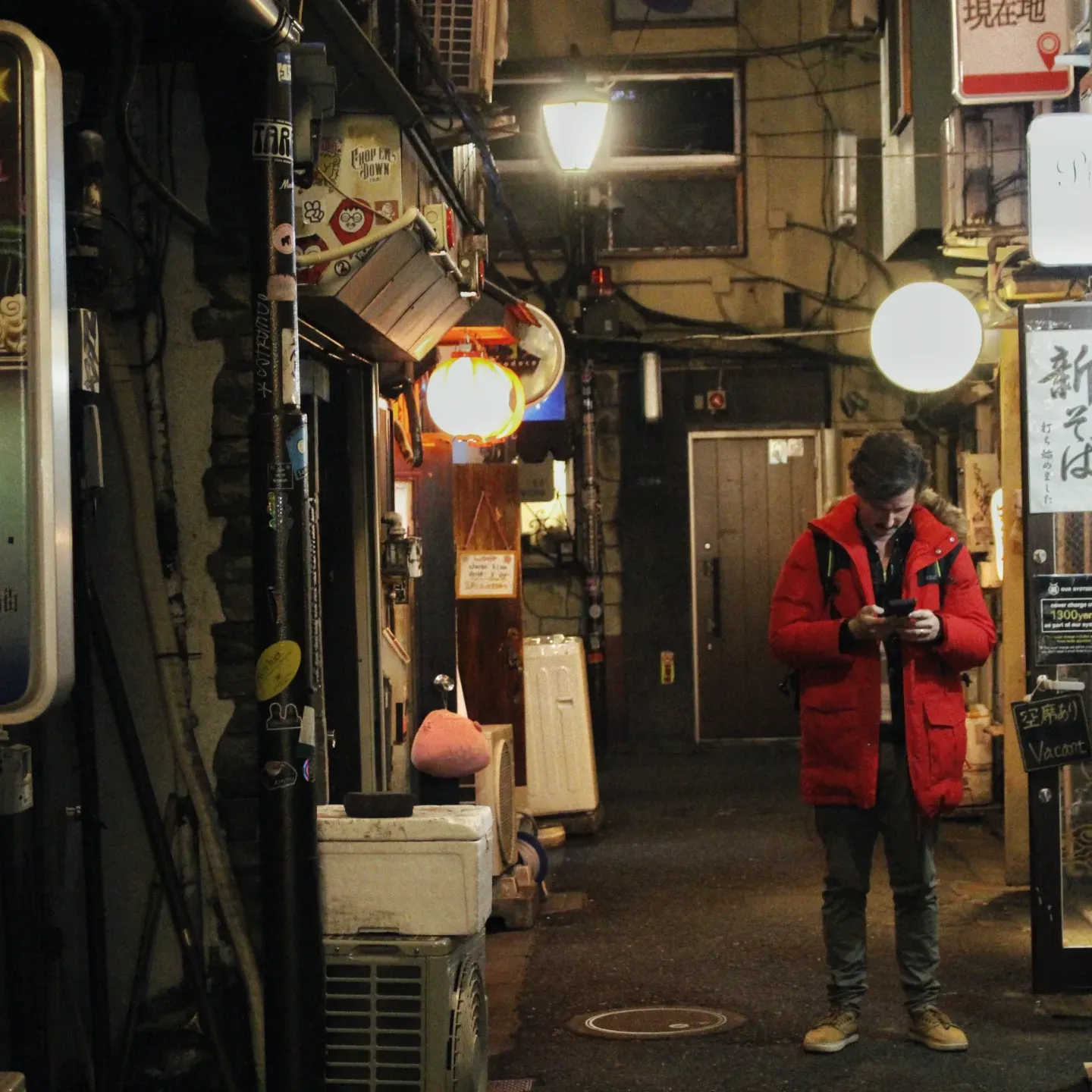 Man in a red jacket checks his phone in a narrow, softly lit Tokyo alley with glowing lanterns at night.