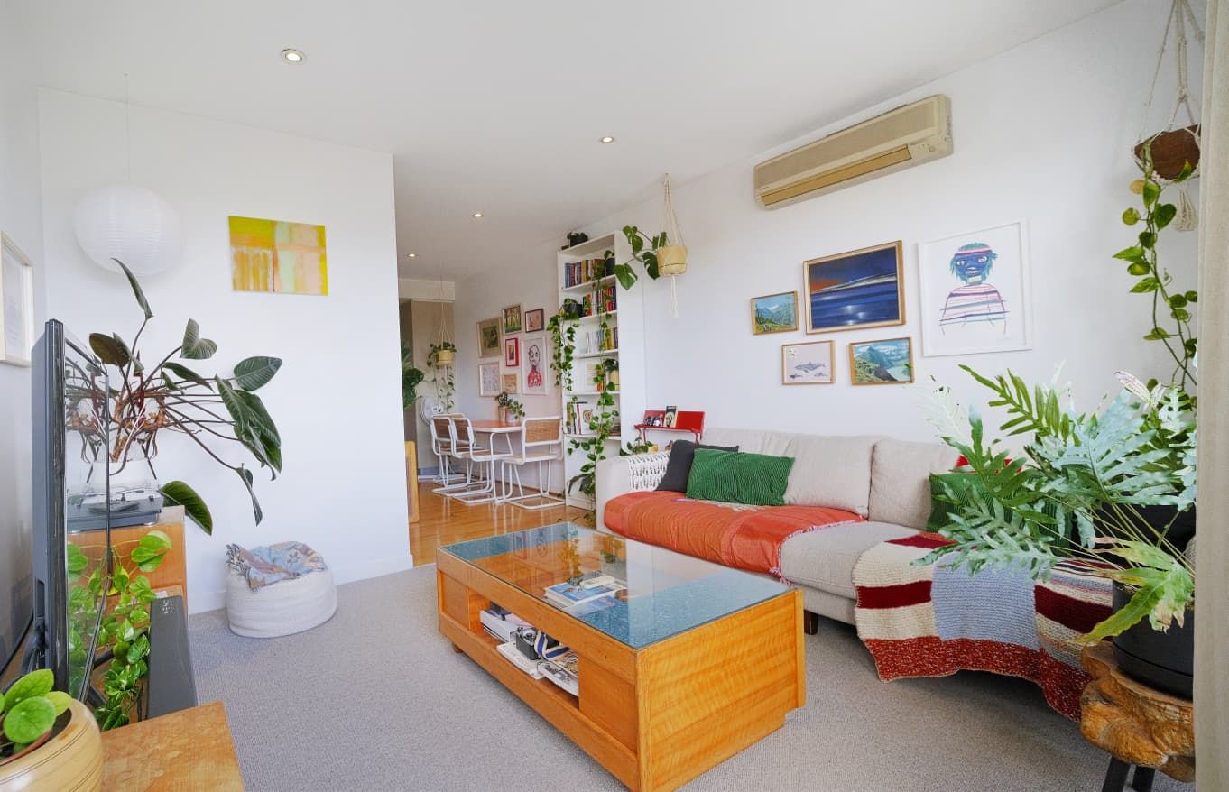 Cozy living room with a beige sofa, wooden coffee table, framed art, wall air conditioner and lush indoor plants.