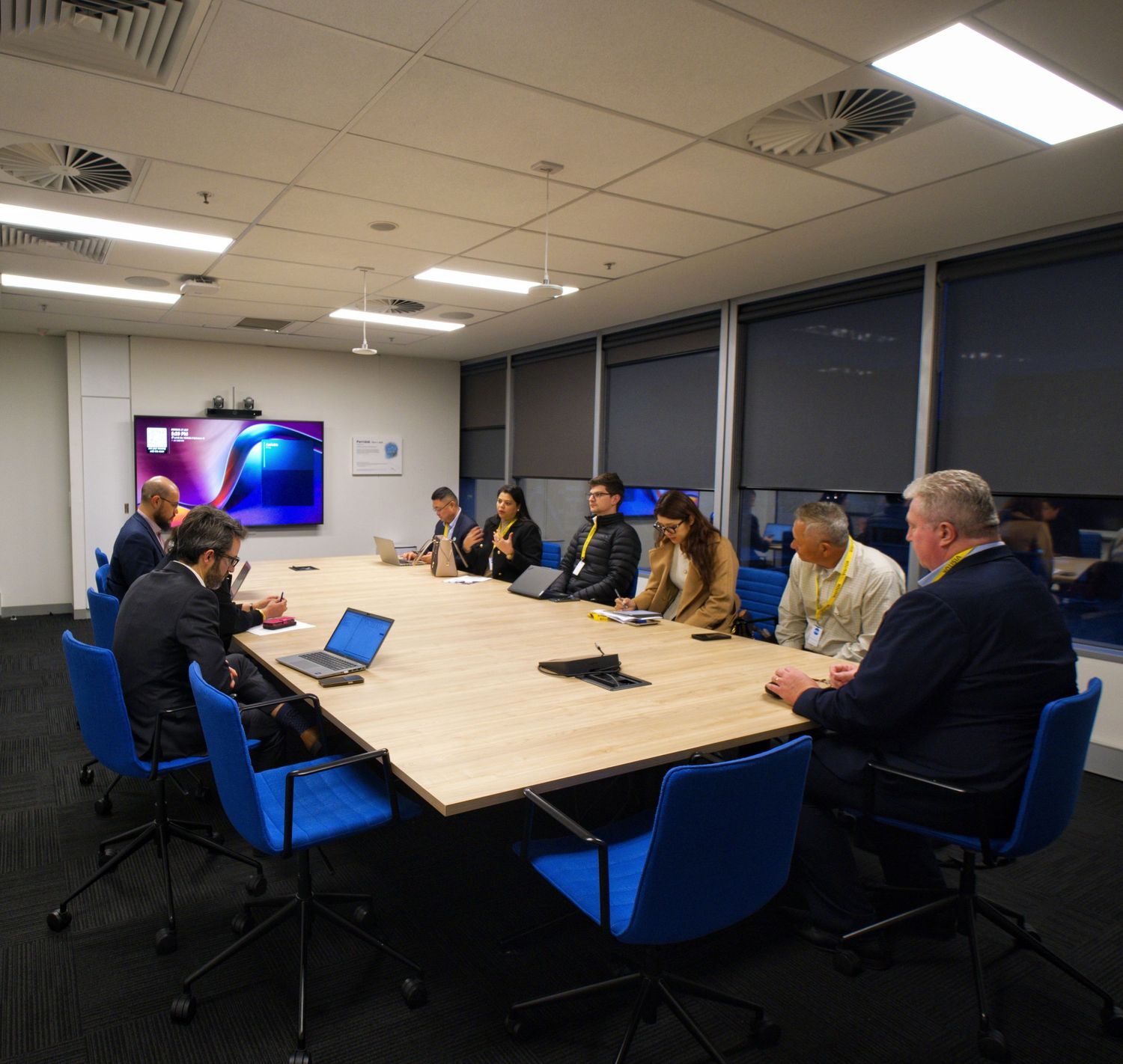Business colleagues gather around a large wooden conference table with blue chairs in a conference room with a wall-mounted screen.