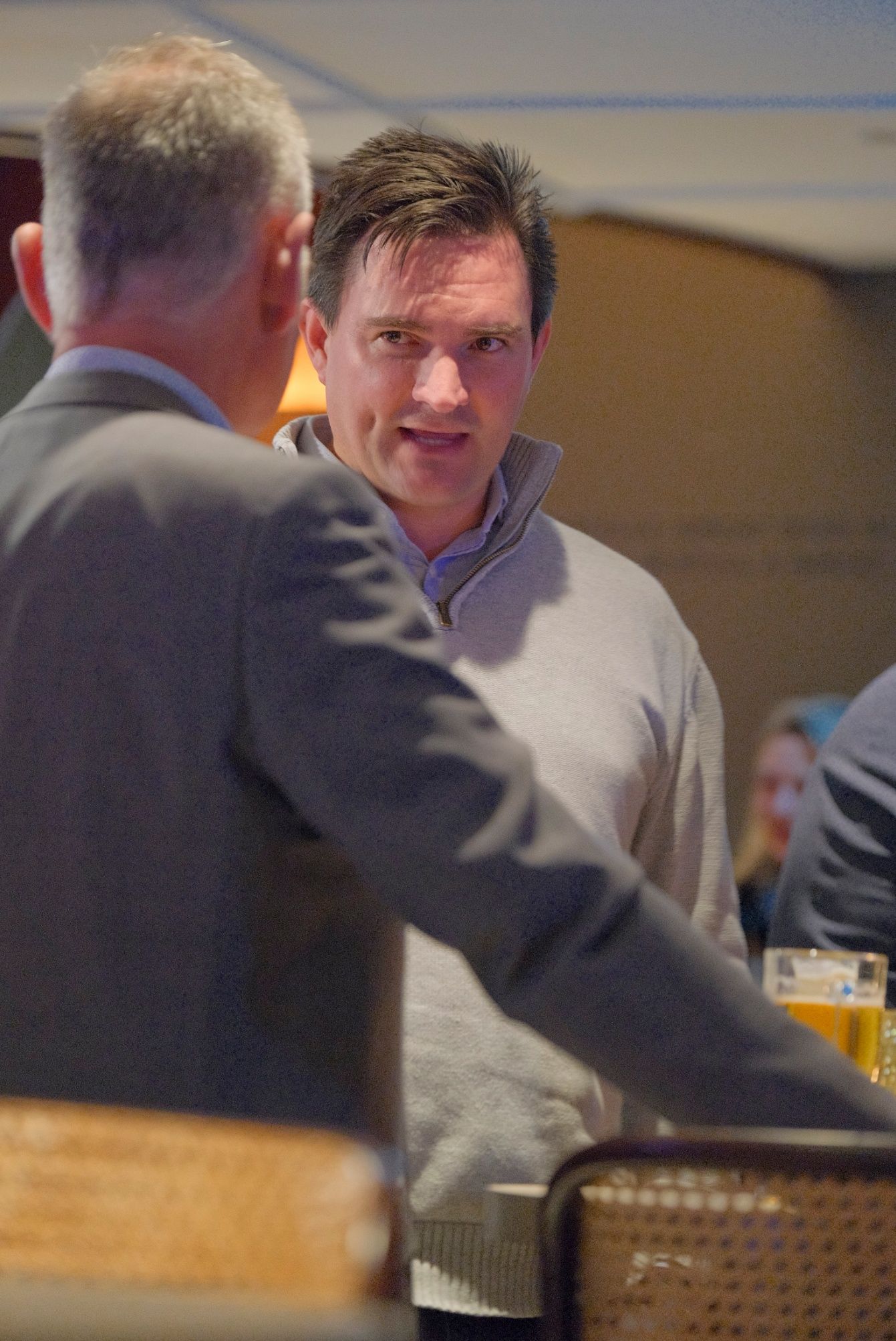 A man in a gray quarter-zip sweater talks with another man at a social event with a beer glass nearby.
