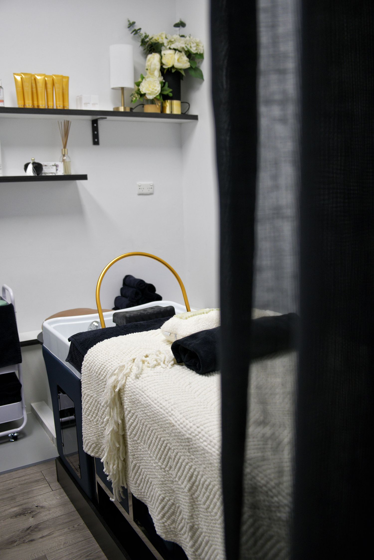 Salon treatment bed with cream knit throw and black towels, framed by a dark curtain, flowers and shelves of gold products.