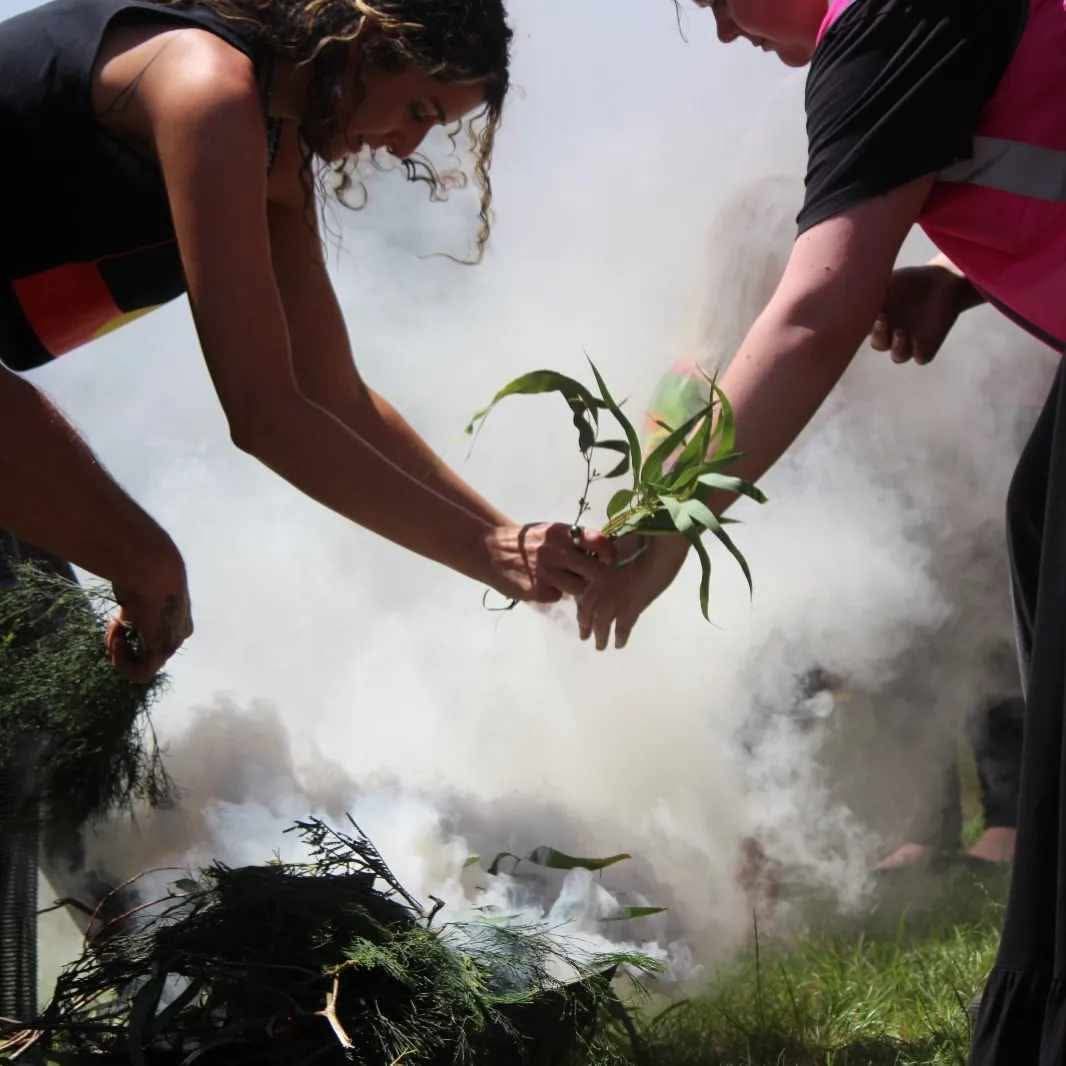 Two people hold green leaves in their hands over a smoking ceremony fire as thick white smoke rises from native plant branches.