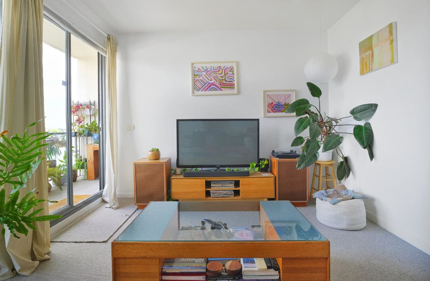 Living area with a wooden TV console, glass-top coffee table, potted plants and a sliding door opening to a balcony.