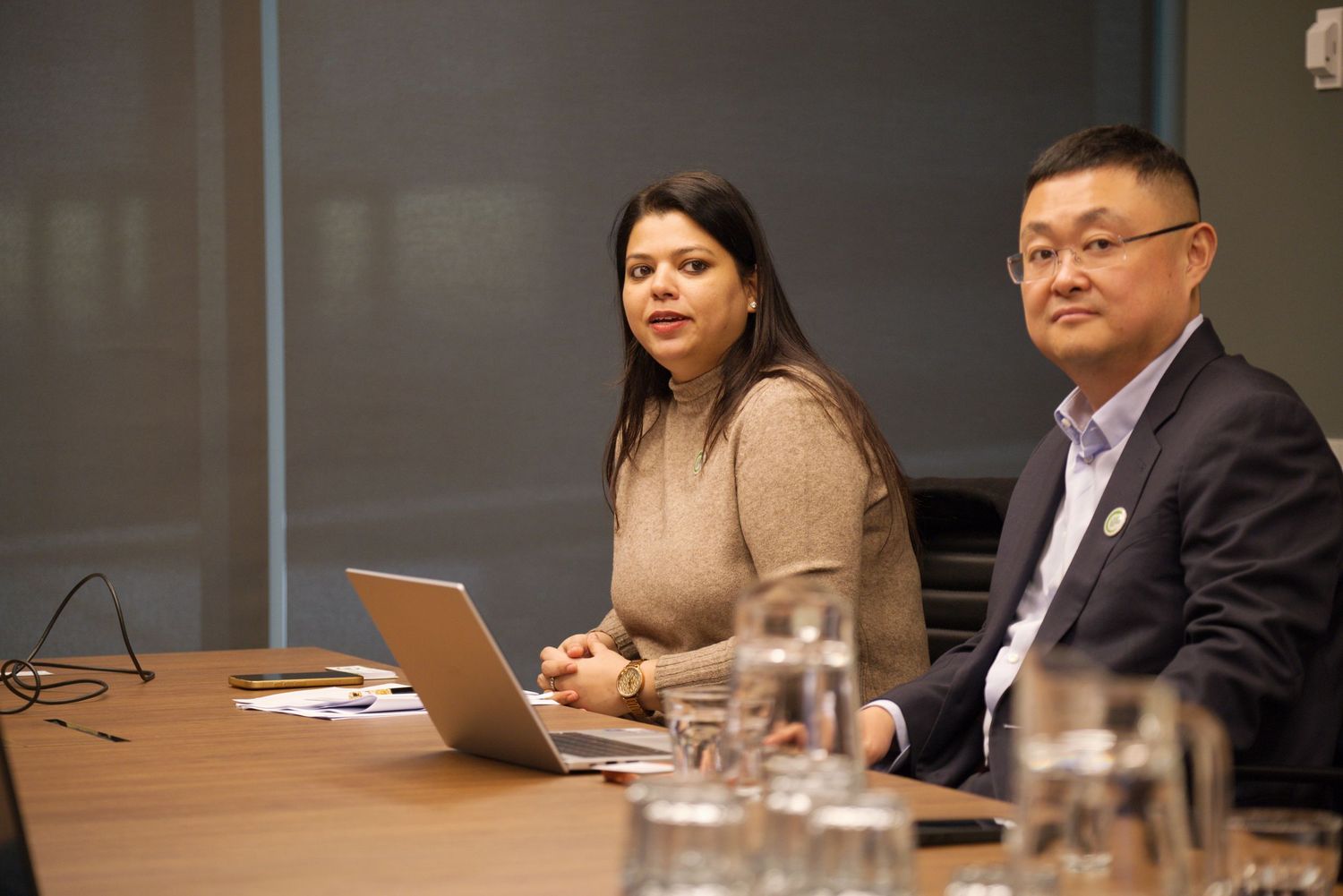 A woman and a man in business attire sit at a conference table with a laptop and glass water pitchers during a meeting.