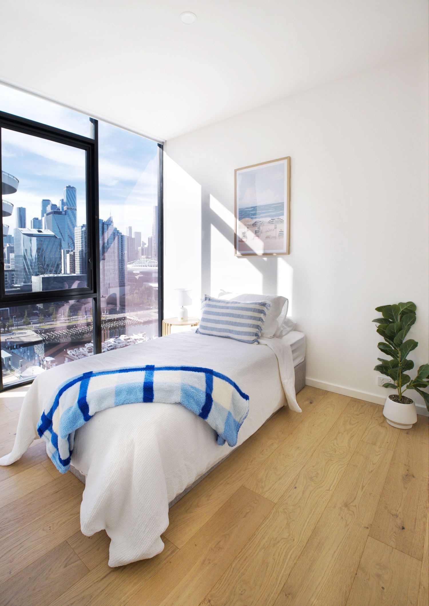 Single bedroom with blue and white bedding, framed beach artwork and floor-to-ceiling windows showing city views.