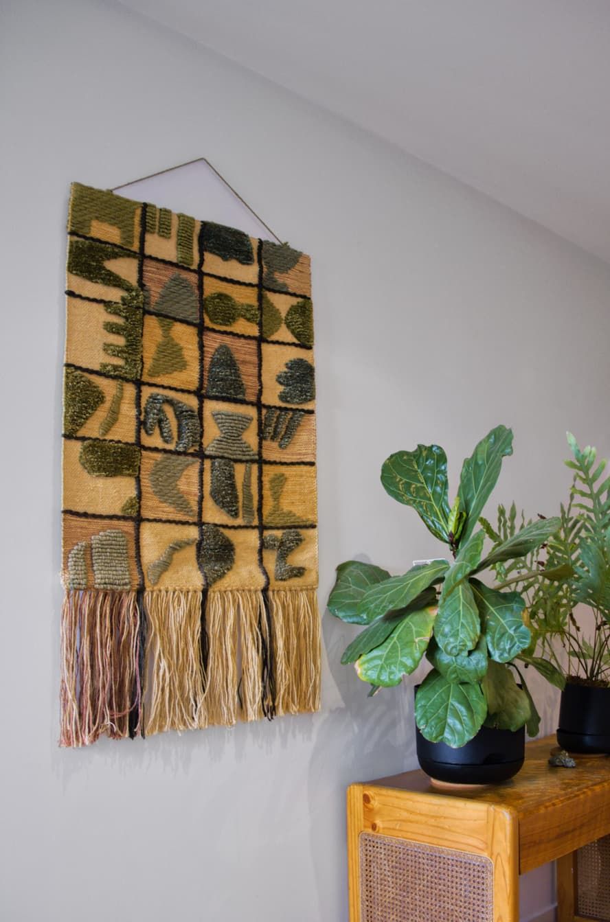 Handwoven wall hanging in earthy green and mustard tones with fringe, beside a fiddle leaf fig on a rattan wooden cabinet.