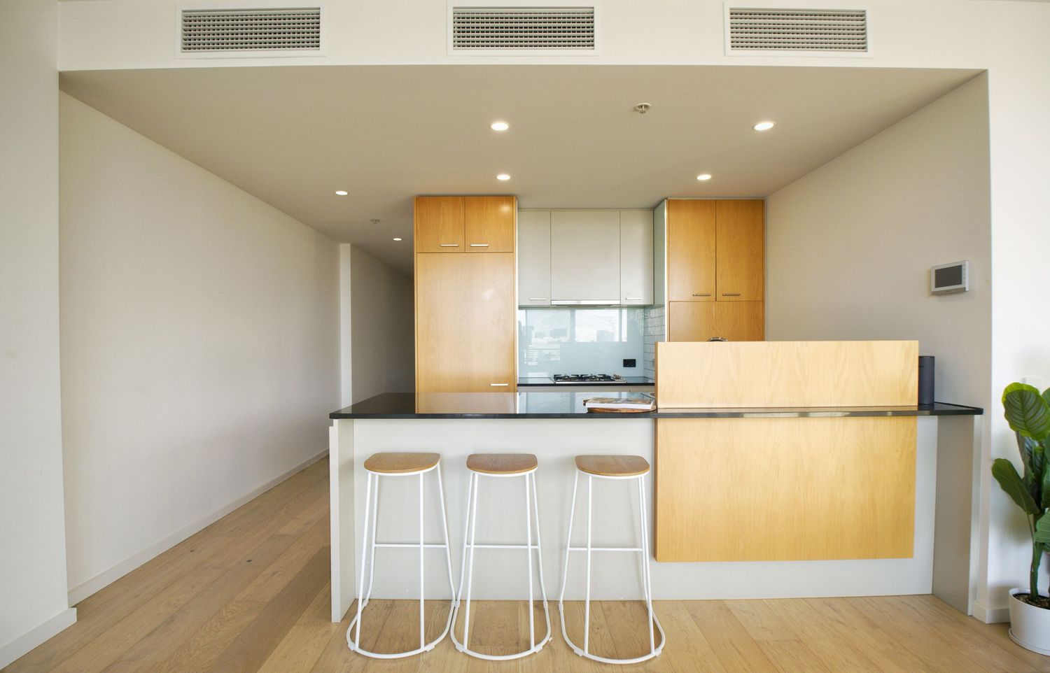 Open kitchen with timber cabinetry, black benchtop, breakfast bar with three stools and ducted air conditioning vents.