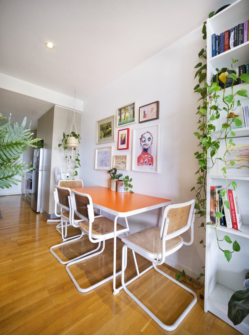 Dining nook with an orange table, white cane-back chairs, a gallery wall of framed art, trailing plants and a bookshelf.