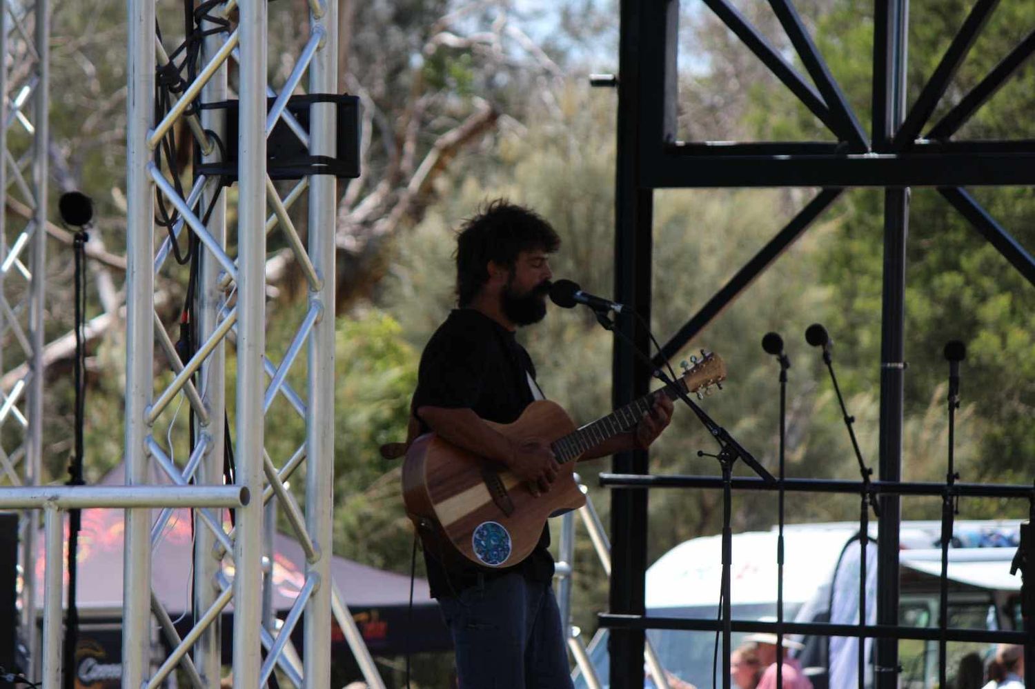 A bearded musician plays an acoustic guitar and sings into a microphone on an outdoor stage framed by metal truss and trees.
