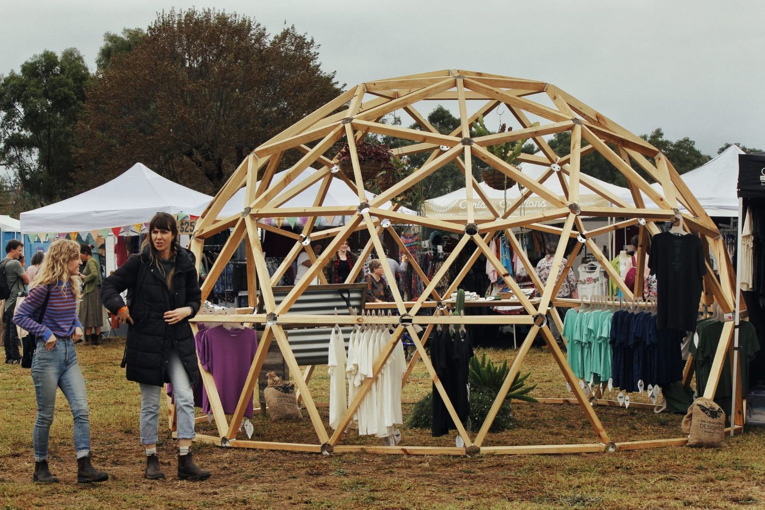 Two women stand in front of a wooden geodesic dome market stall displaying hanging clothing at an outdoor festival.