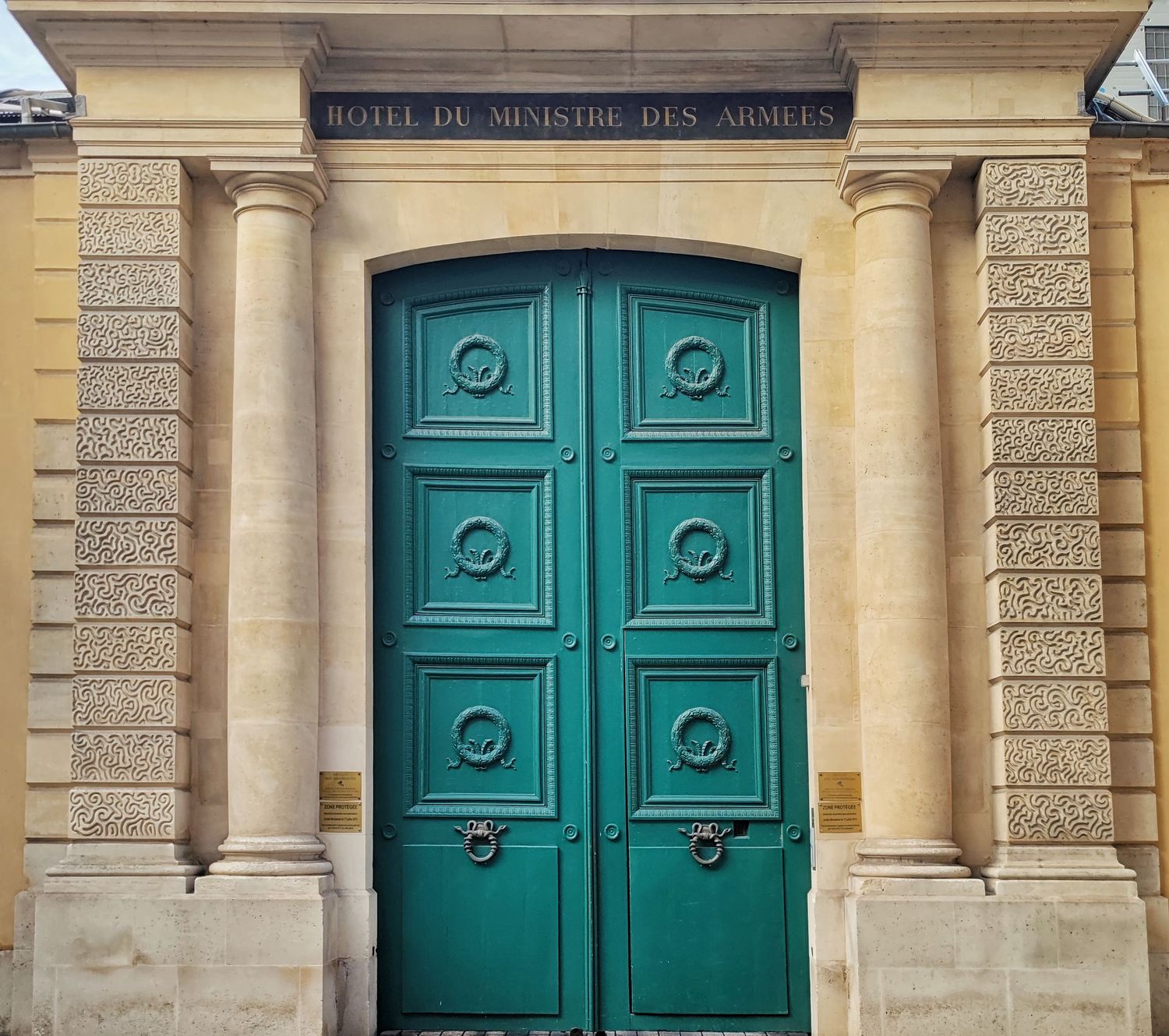 Teal double door with decorative panels and ring handles beneath the Hôtel du Ministre des Armées inscription between stone columns.