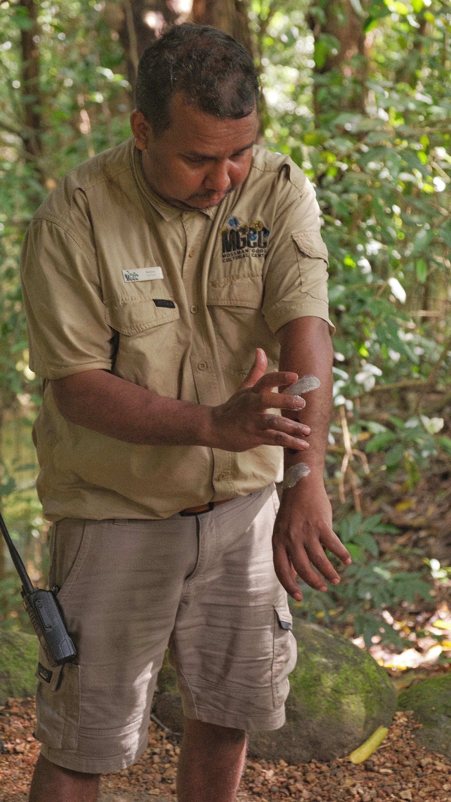 A park ranger in a tan uniform demonstrates a bush technique in a lush green rainforest, with a radio clipped to his belt.